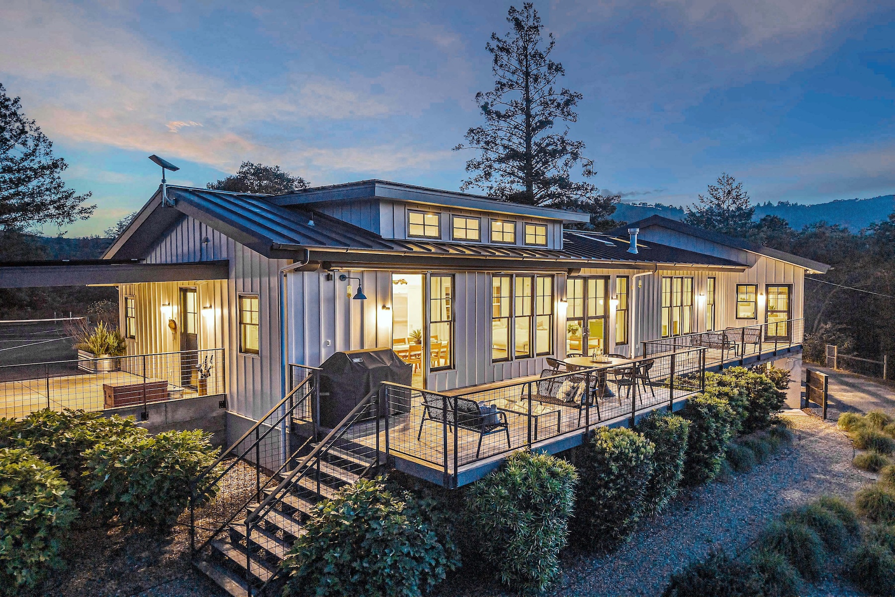 This elevated rear view captures a modern farmhouse-style home at dusk, illuminated by warm interior and exterior lighting. The structure features vertical siding, a metal roof, and a spacious deck with black metal railings, perfect for outdoor dining and relaxation. The perspective highlights the home's integration into the natural landscape, with a gentle slope leading down to a gravel path and manicured shrubbery.