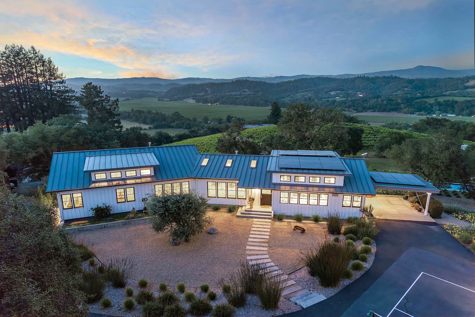This stunning aerial perspective captures a modern farmhouse-style residence nestled in a lush, rolling landscape at twilight. The home features a distinctive blue metal roof, white siding, and numerous windows that glow warmly against the soft evening sky. A stone-paved walkway leads through a landscaped gravel courtyard, while the surrounding property showcases expansive vineyards and distant mountain vistas.