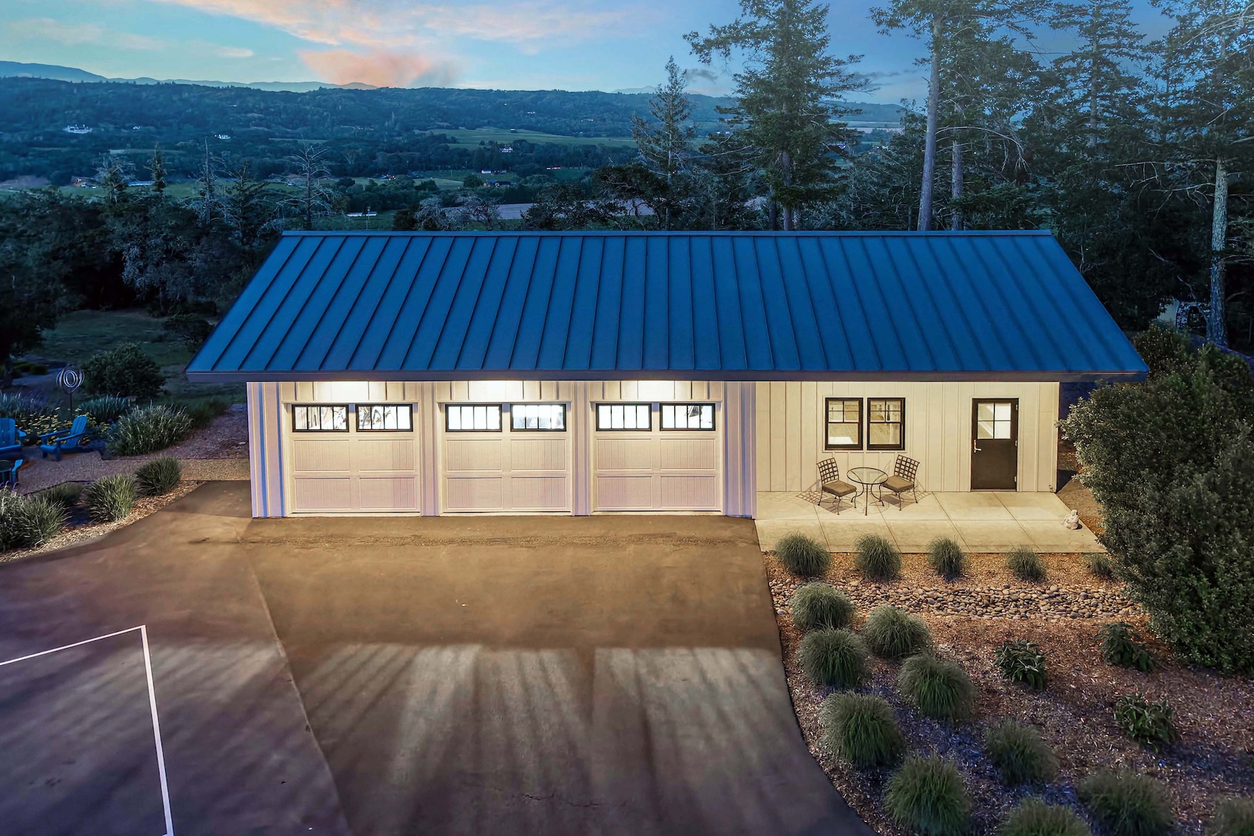 This aerial perspective captures a modern, detached three-car garage featuring a striking blue metal roof and light-colored vertical siding. The structure includes a small, covered patio area with bistro seating, illuminated by warm interior lighting that contrasts with the twilight sky. The surrounding landscape is neatly manicured with ornamental grasses and stone accents, set against a backdrop of mature trees and distant rolling hills.
