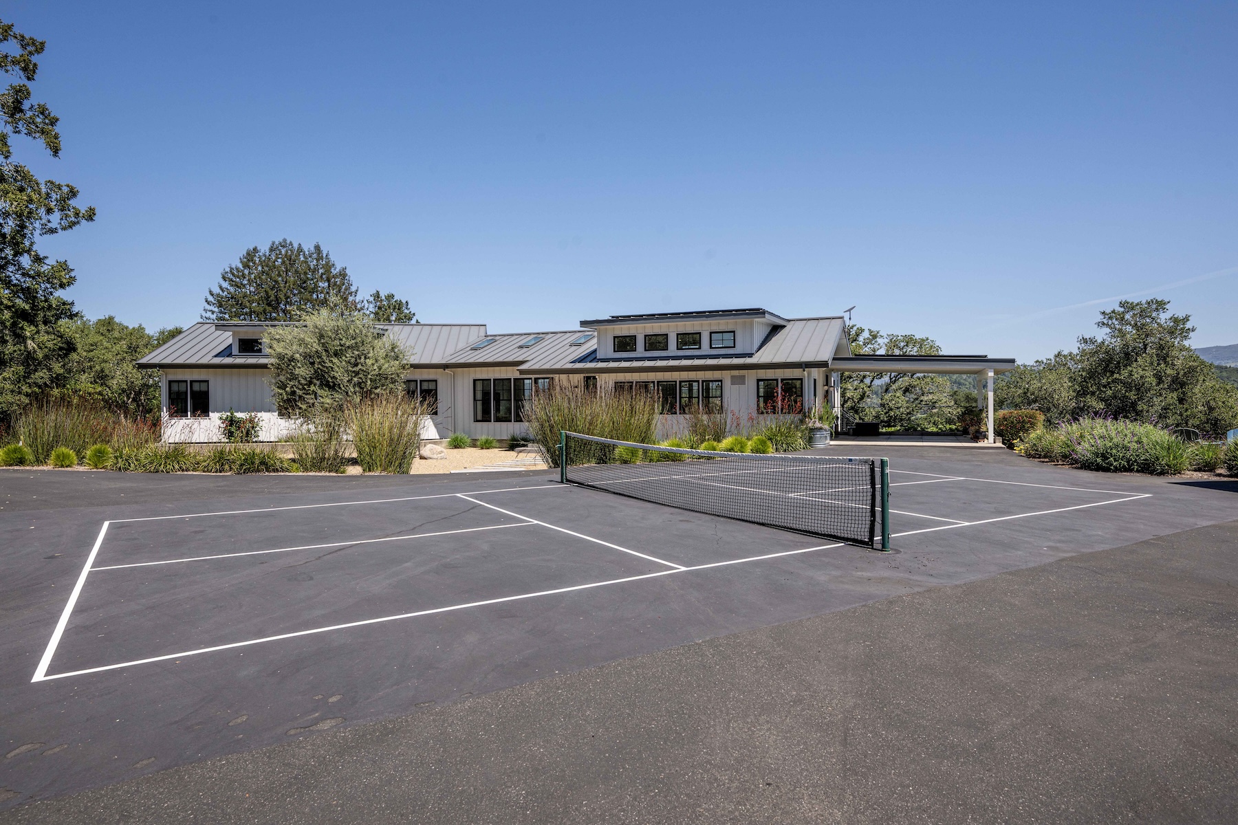 This exterior view showcases a modern, single-story home with a metal roof and white siding, set against a clear blue sky. In the foreground, a paved area serves as a multi-purpose court with a tennis net, while the surrounding landscape features native grasses and mature trees. The perspective is wide and eye-level, emphasizing the spaciousness of the property and its integration with the natural environment.
