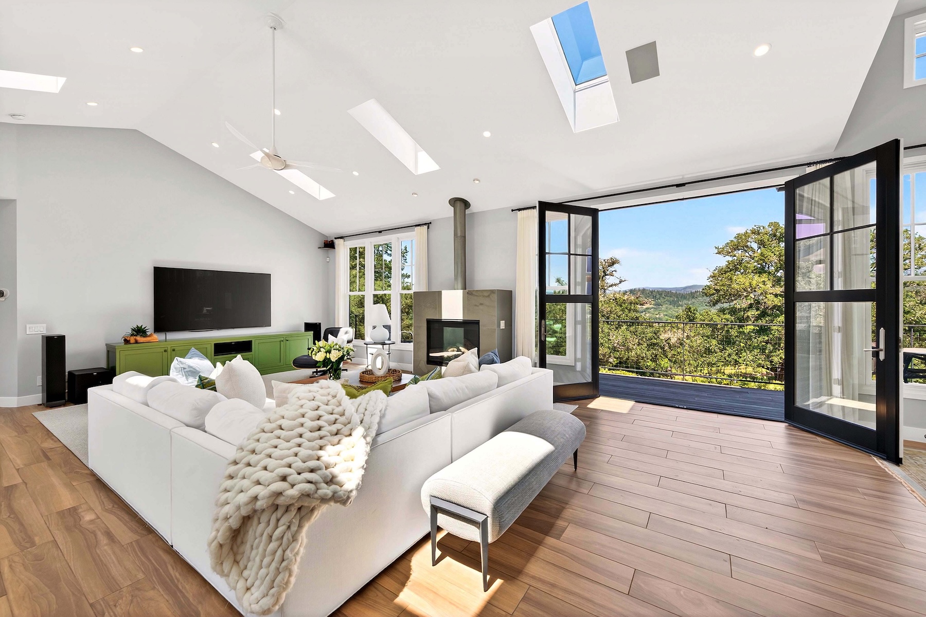 This bright and airy living room features a vaulted ceiling with multiple skylights, creating an expansive and open atmosphere. A large white sectional sofa with a chunky knit throw blanket serves as the centerpiece, positioned in front of a television and a tall, stone-clad chimney breast. The space seamlessly transitions to an outdoor deck through wide-opening glass doors, offering panoramic views of the surrounding landscape.