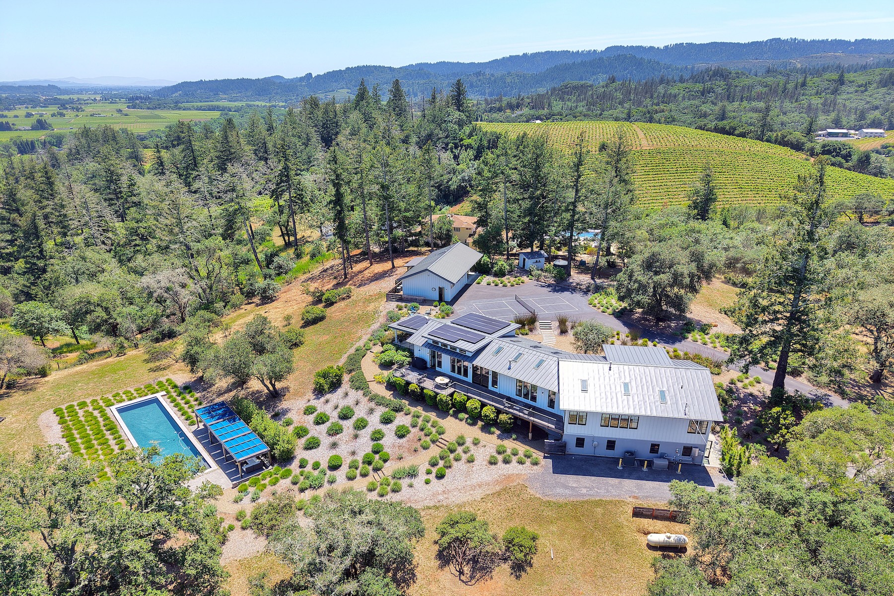 This high-angle aerial view captures a sprawling rural estate nestled among rolling hills and vineyards. The property features a modern, multi-sectioned residence with a metal roof and solar panels, a rectangular swimming pool with an adjacent shaded structure, and a detached garage building with a paved court. The surrounding landscape is characterized by mature trees, native plantings, and expansive natural terrain, offering a serene and private atmosphere.