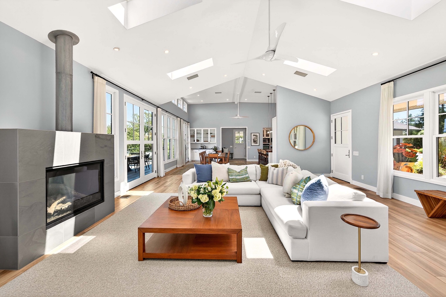 This bright and airy living room features a vaulted ceiling with skylights and a large, comfortable sectional sofa arranged around a central wood coffee table. A prominent stone-clad chimney breast anchors the space, while light-toned wood flooring and soft blue wall paint create a serene, coastal-inspired atmosphere. The open-concept layout provides a clear view through to the dining area and kitchen, emphasizing the home's spacious and inviting design.