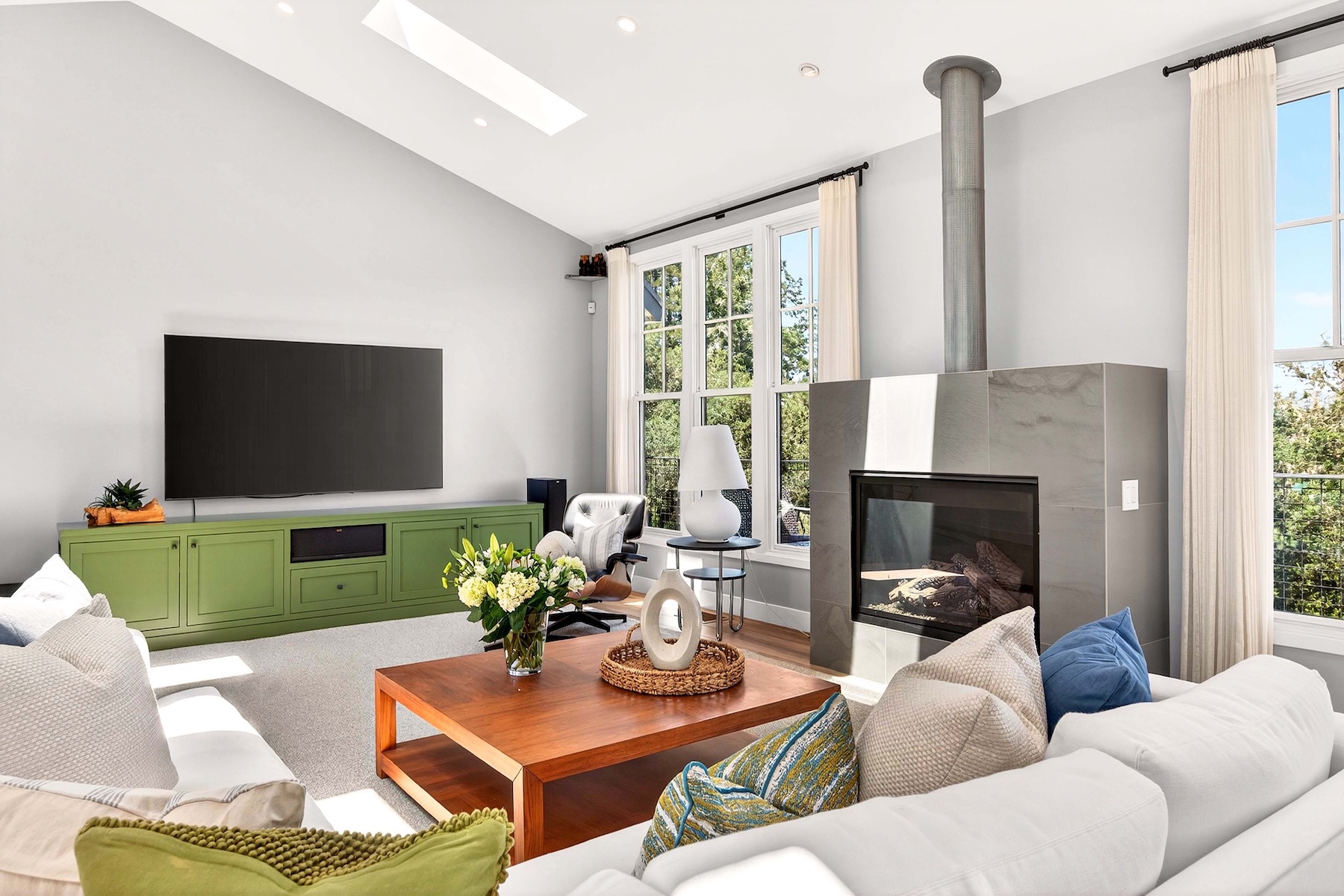 This bright and airy living room features a vaulted ceiling with a skylight and a prominent fireplace with a tall, gray stone-clad chimney breast. A large, olive-green media console sits beneath a wall-mounted television, while a wooden coffee table serves as the centerpiece for the seating area. The space is framed by tall windows with white curtains, creating a welcoming atmosphere that blends modern design with comfortable living.