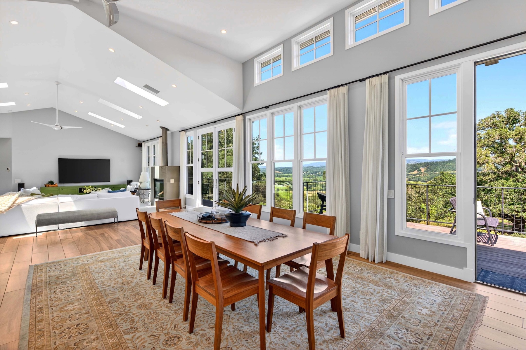 This bright and airy dining area features a long wooden table surrounded by eight matching chairs, set atop a large, intricately patterned area rug. The room is defined by its vaulted ceiling with skylights and a wall of tall windows that provide abundant natural light and expansive views of the surrounding landscape. Adjacent to the dining space, a living area with a white sectional sofa and a fireplace creates an open-concept layout perfect for entertaining.