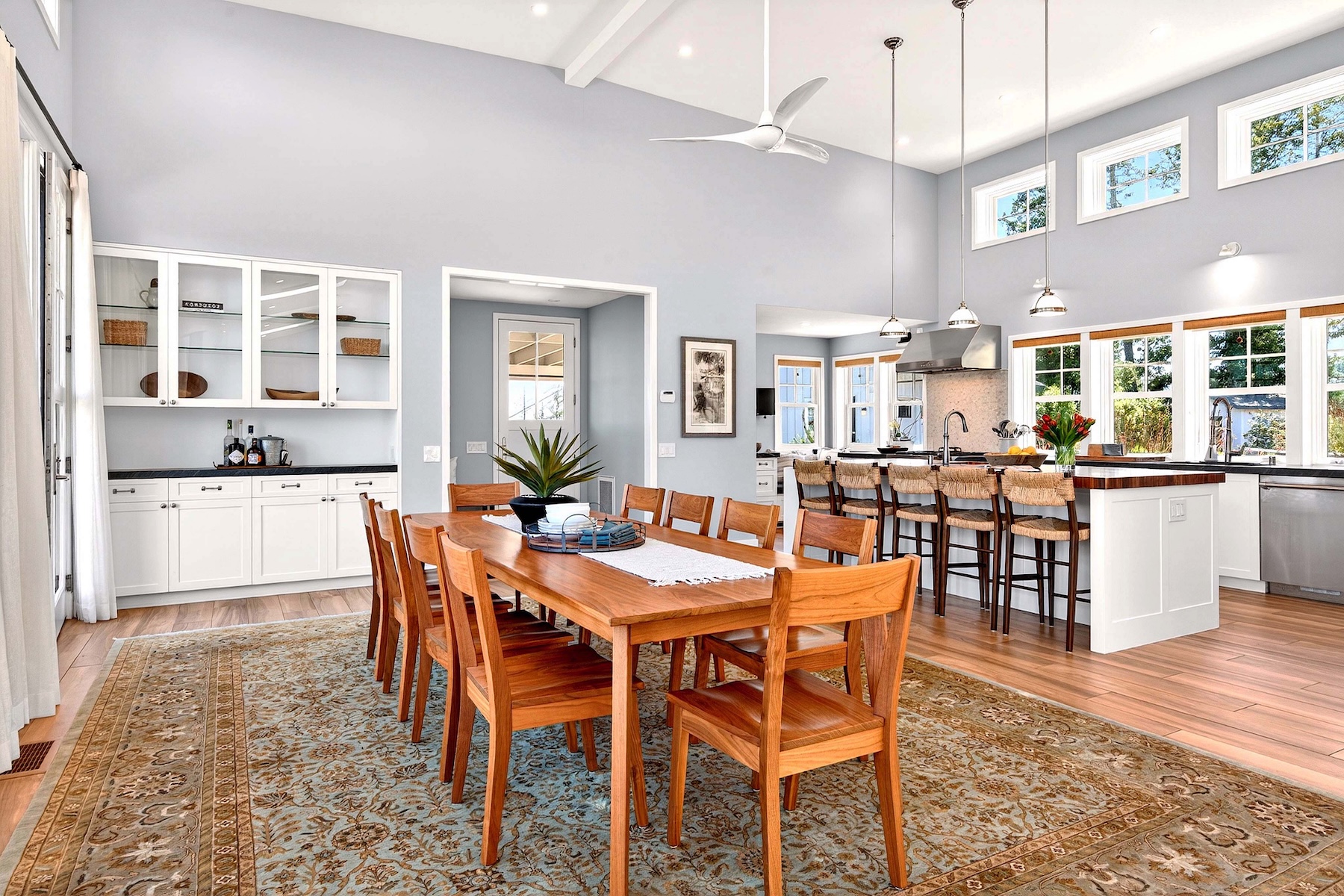This open-concept dining area features a large wooden table surrounded by matching chairs, set atop a detailed patterned rug. The space flows seamlessly into a bright kitchen with a breakfast bar, white cabinetry, and pendant lighting. The room is characterized by high ceilings, light-toned walls, and large windows that provide ample natural light, creating an airy and inviting atmosphere.