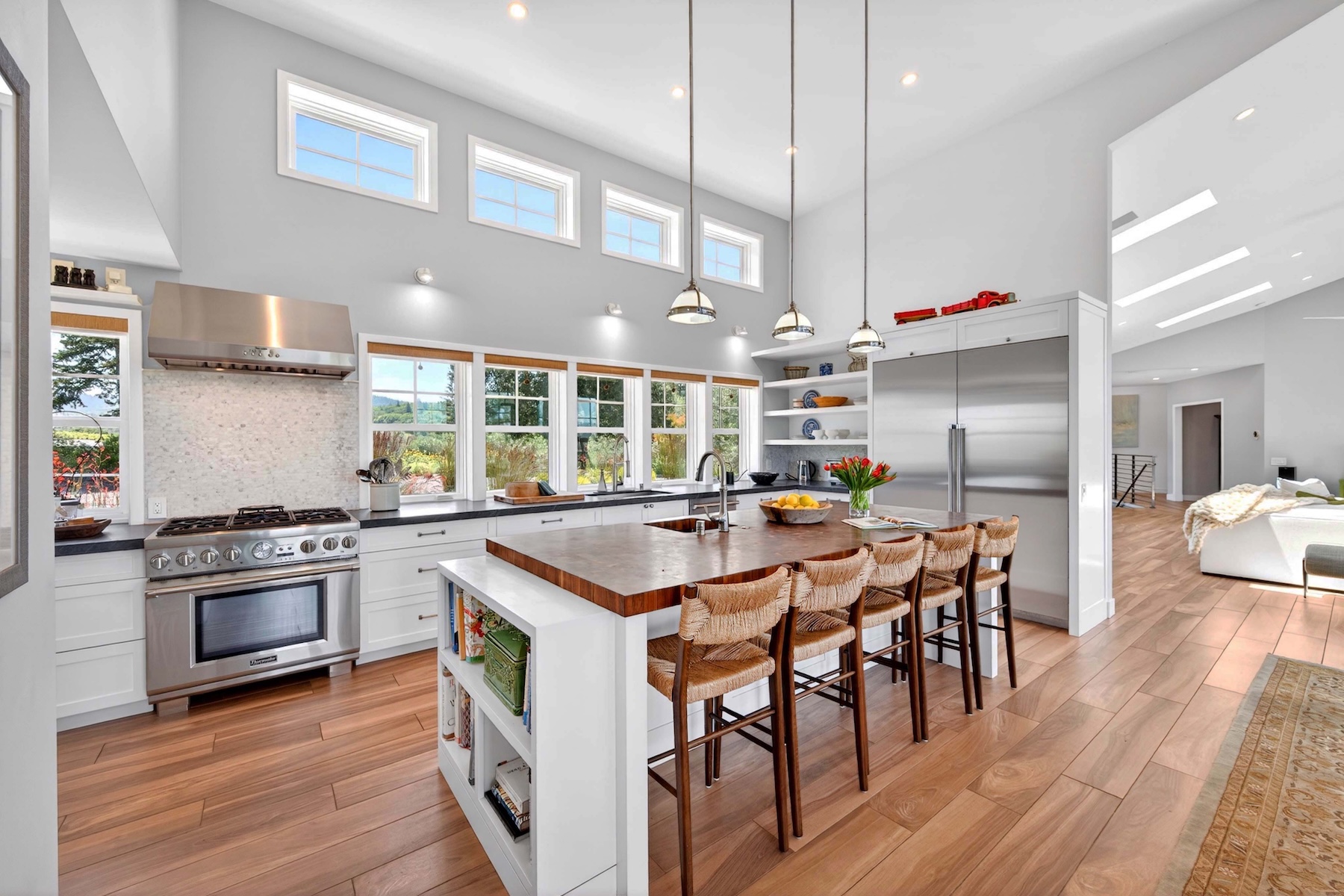 This bright and airy kitchen features a large central island with a wood-toned countertop and woven bar stools, serving as the focal point of the space. White cabinetry is paired with stainless steel appliances, including a professional-grade range and a double-door refrigerator, while a row of windows above the counter brings in abundant natural light. The room is finished with light wood flooring and elegant pendant lighting, creating a warm and inviting atmosphere that flows into an adjacent living area.