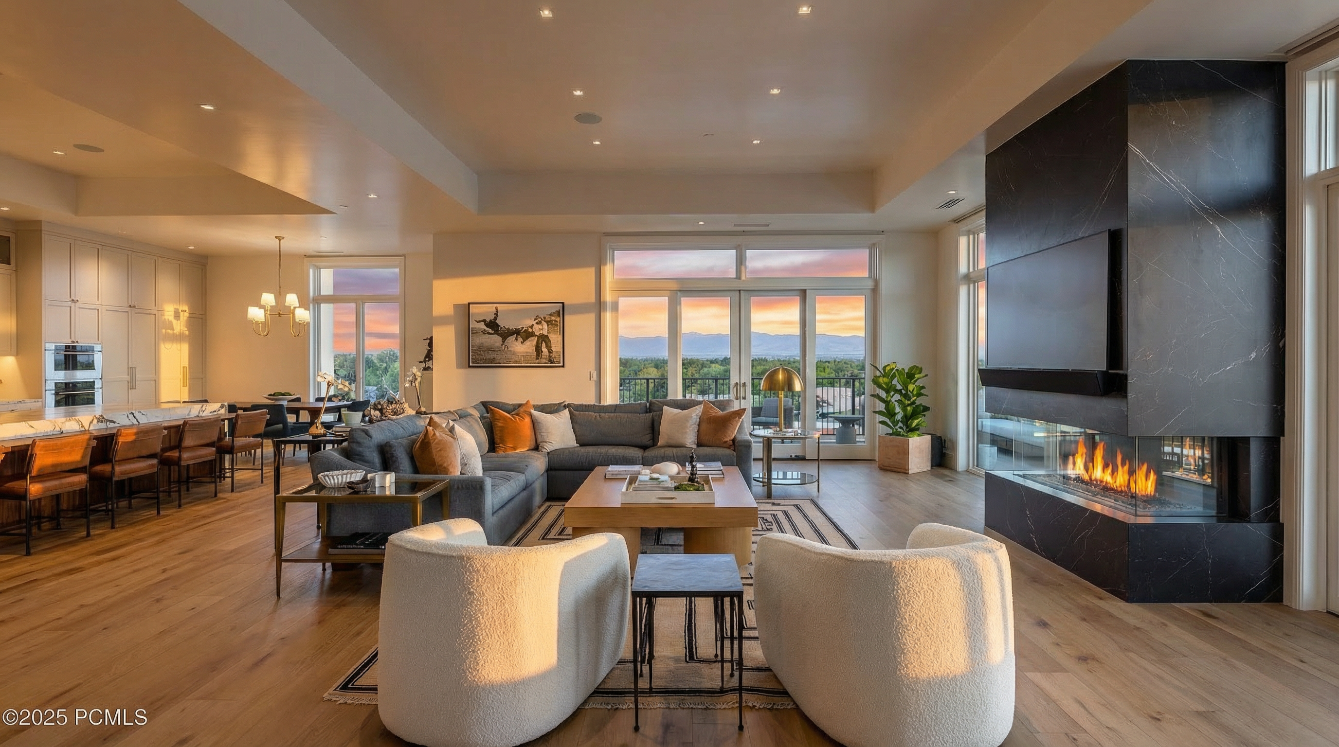 This is a well-lit, inviting living room featuring hardwood floors, a modern fireplace, and neutral-toned furniture. The open-concept design flows seamlessly into the kitchen and dining area, creating a spacious and airy feel. Large windows offer scenic views, enhancing the room's overall appeal.