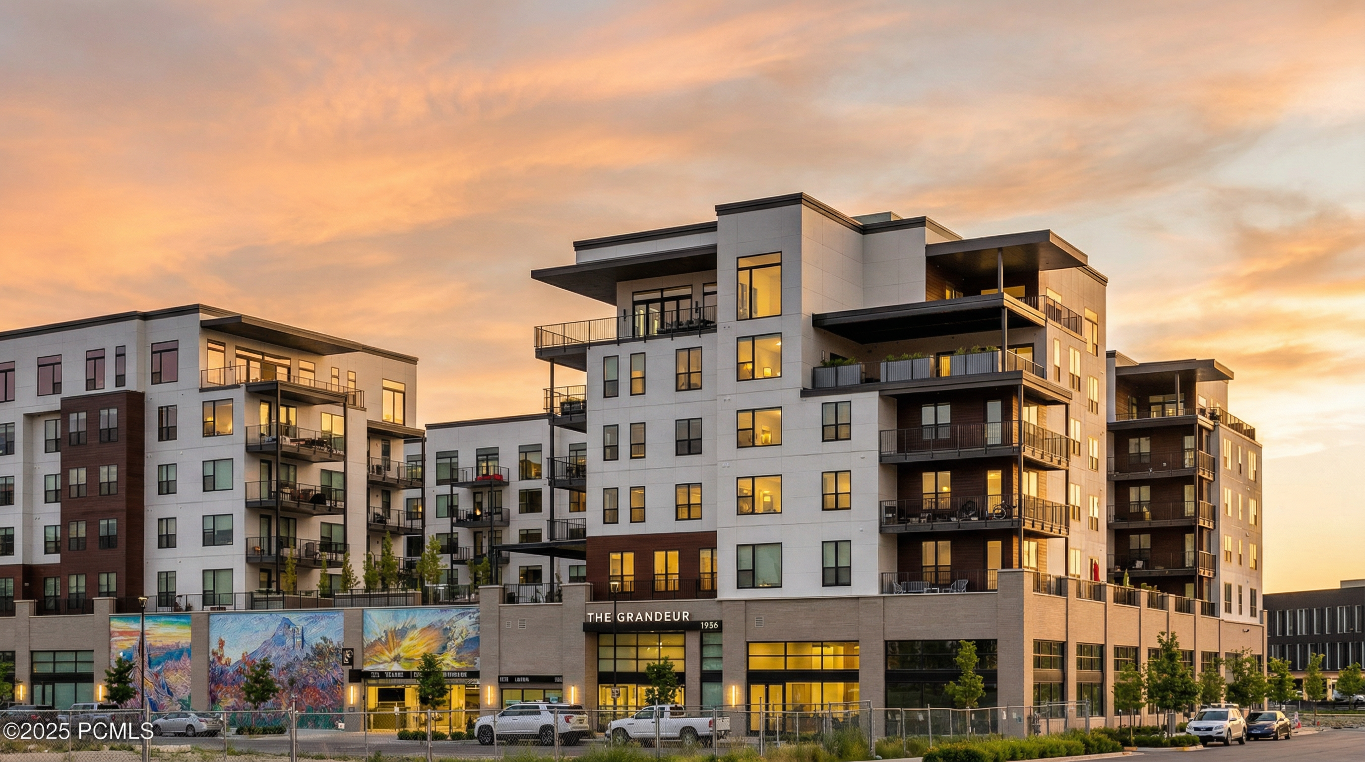 This is a front view of a modern apartment complex, featuring clean lines, a mix of white and brown paneling, and multiple balconies. The building is named 'The Grandeur' and appears to be well-maintained. The colorful mural along the base adds an artistic element.