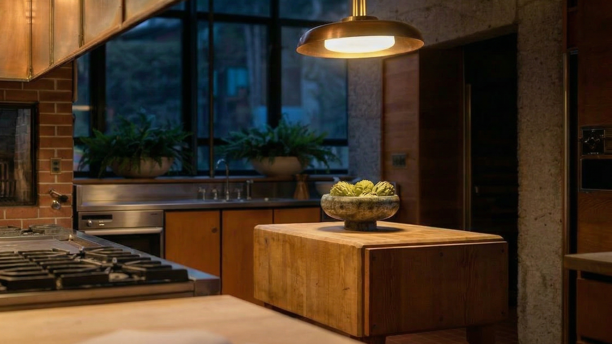 This warm and inviting kitchen space features a distinctive wooden butcher block island as a centerpiece, topped with a bowl of fresh artichokes. The design includes natural wood cabinetry, a stainless steel gas range, and warm under-cabinet lighting, all set against a textured brick wall. Above, a sleek, modern pendant light casts a soft glow, creating a sophisticated and cozy atmosphere.