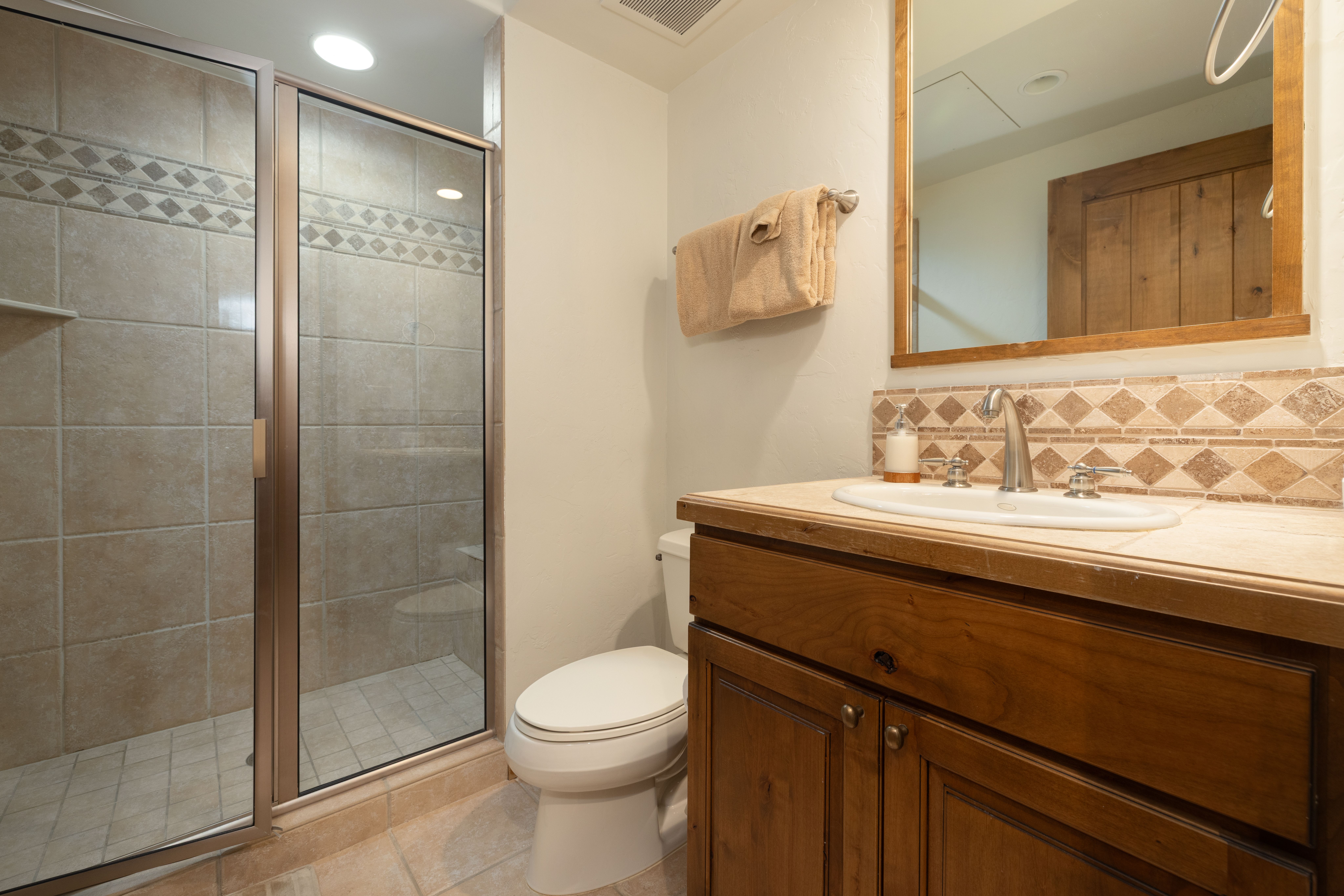 This bathroom features a rustic wooden vanity with a stone countertop and a decorative tile backsplash that adds texture and warmth. It includes a walk-in shower with a glass enclosure and matching tiled walls, alongside a standard white toilet. The space provides a functional and well-maintained look suitable for a guest or secondary bathroom.