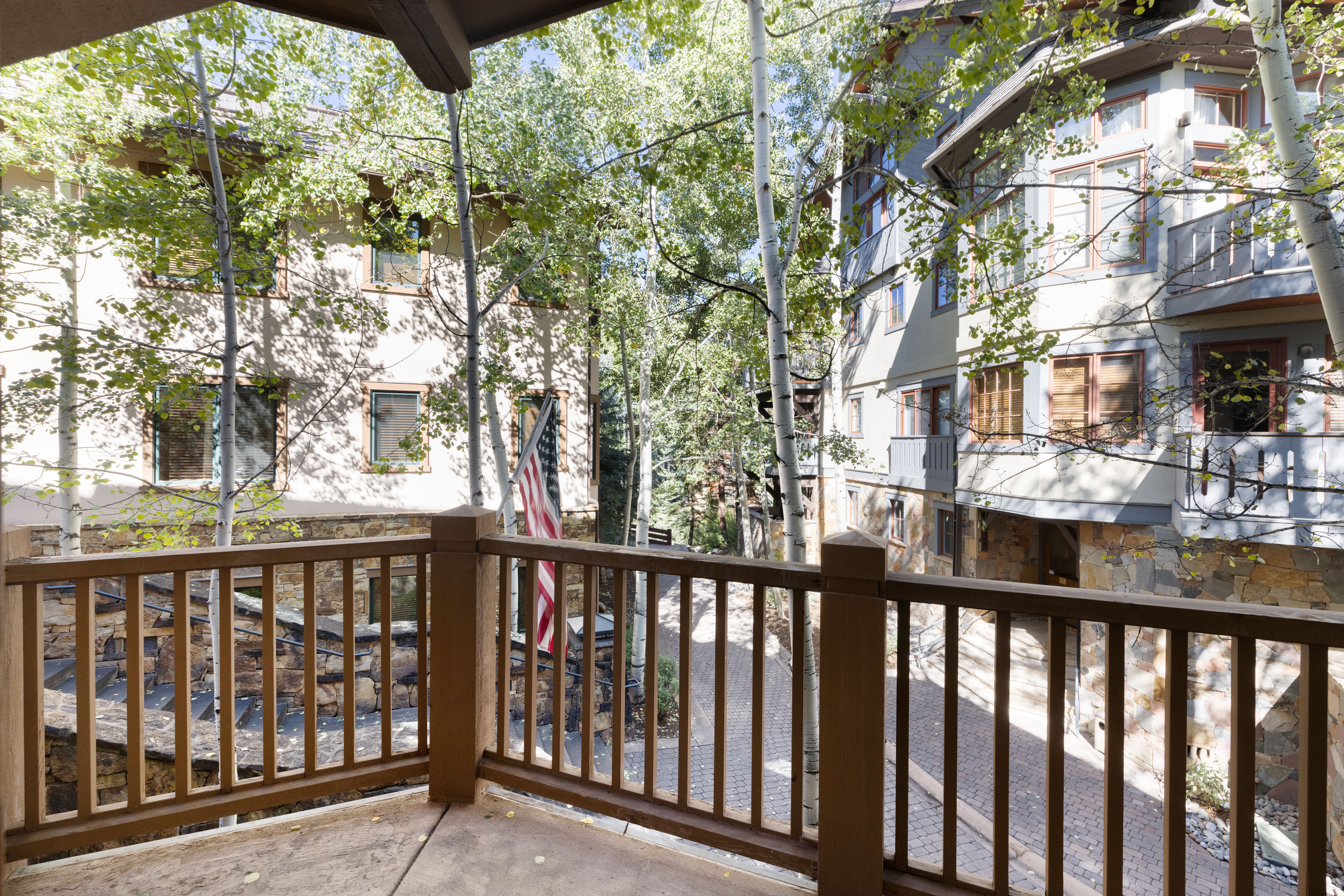 This view is captured from an elevated wooden deck, providing a scenic perspective of the surrounding complex architecture and natural landscape. The space features sturdy wooden railings and overlooks a paved pedestrian walkway flanked by tall birch trees and stone-faced building facades. The setting feels peaceful and well-integrated into a wooded environment, typical of a mountain resort or vacation property.