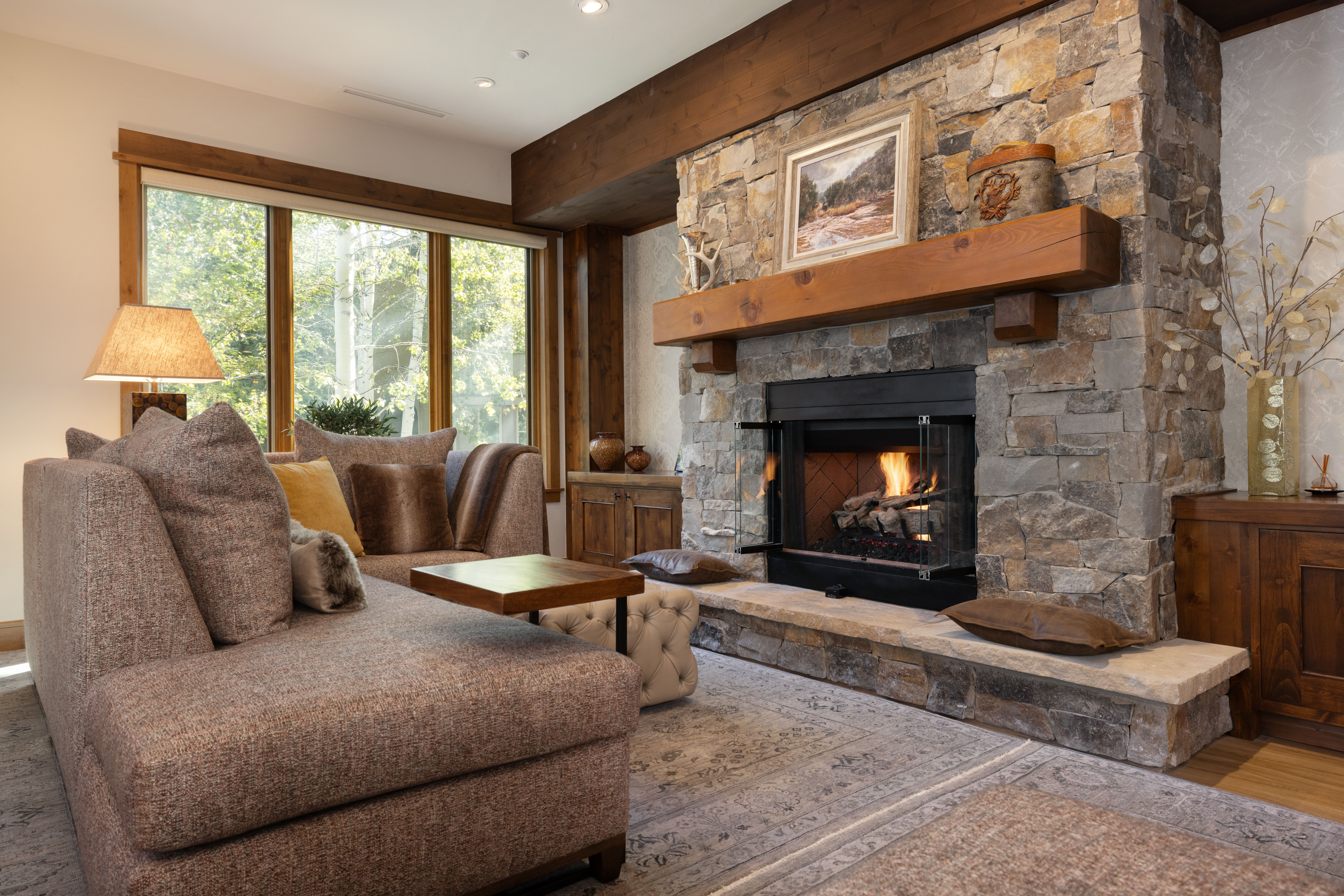 This cozy living room features a striking stone fireplace with a rustic wood mantel as its focal point, complemented by warm wood-toned ceiling beams. A plush, textured sectional sofa paired with an ornate rug creates an inviting seating area, while large windows bring in ample natural light. The overall aesthetic is warm and sophisticated, embodying a luxury mountain lodge style.