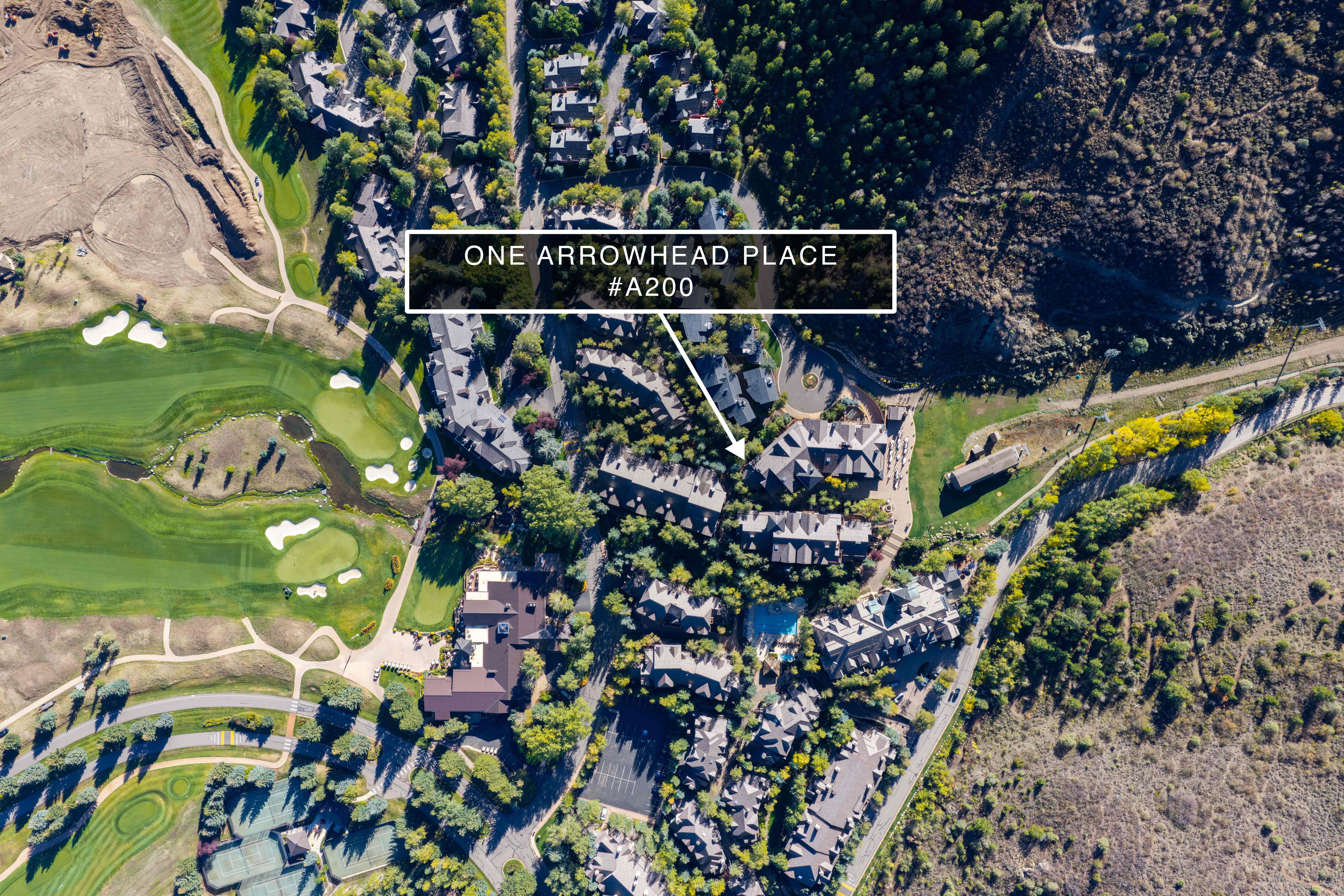 This aerial view showcases a residential property complex integrated seamlessly into a lush, mountainous landscape alongside a golf course. The image highlights the building's location with a clear indicator arrow pointing to One Arrowhead Place #A200, surrounded by mature trees and paved pathways. The setting offers a serene, resort-style atmosphere with convenient access to recreational facilities.