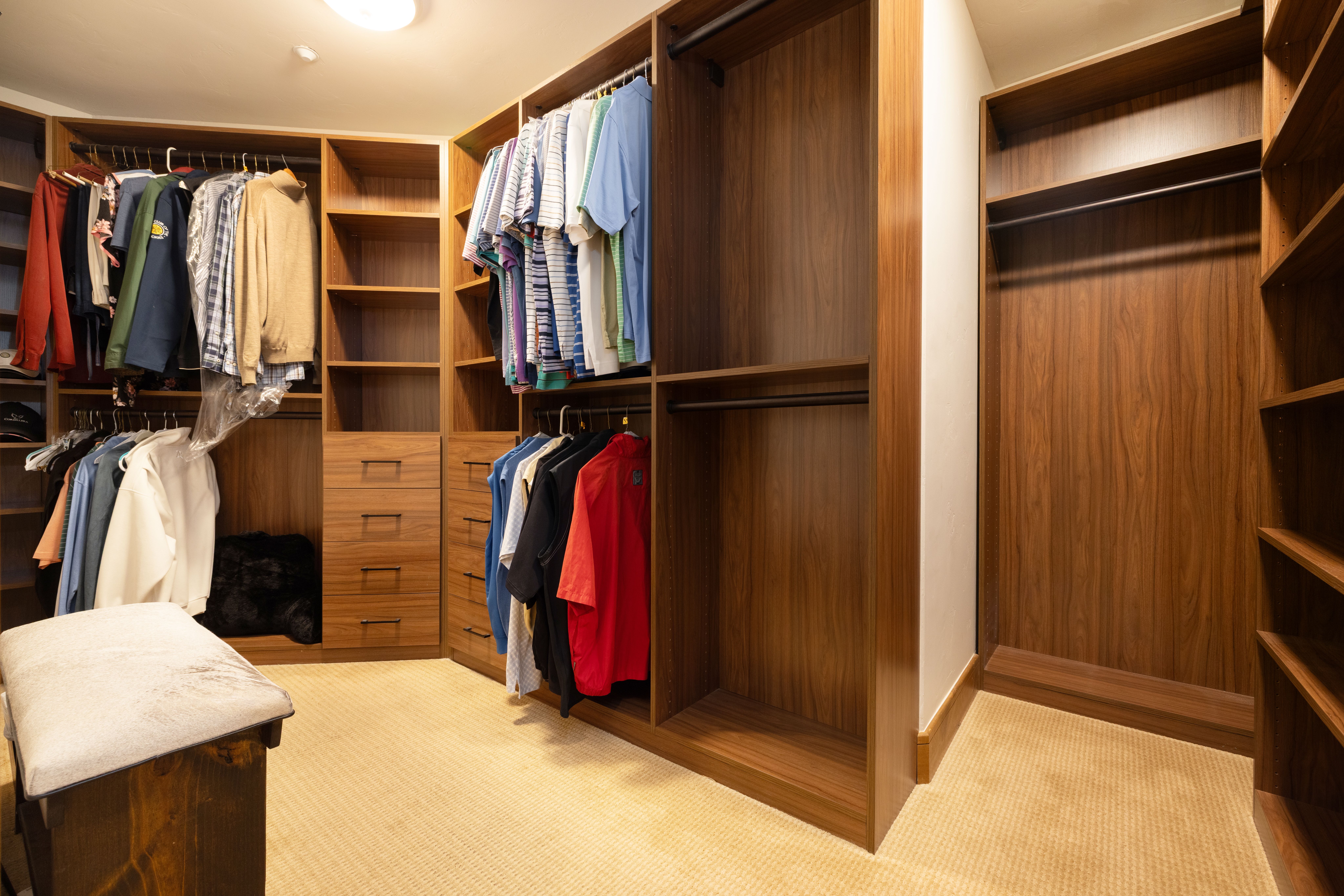 This spacious, custom-designed walk-in closet features rich dark-wood cabinetry, ample hanging space, and integrated shelving with drawers for organized storage. The room is outfitted with high-quality woodwork, neutral wall-to-wall carpeting, and a central ottoman, providing a sophisticated and functional storage solution. Its organized layout and premium finishes make it a highly desirable feature for a luxury residential property.