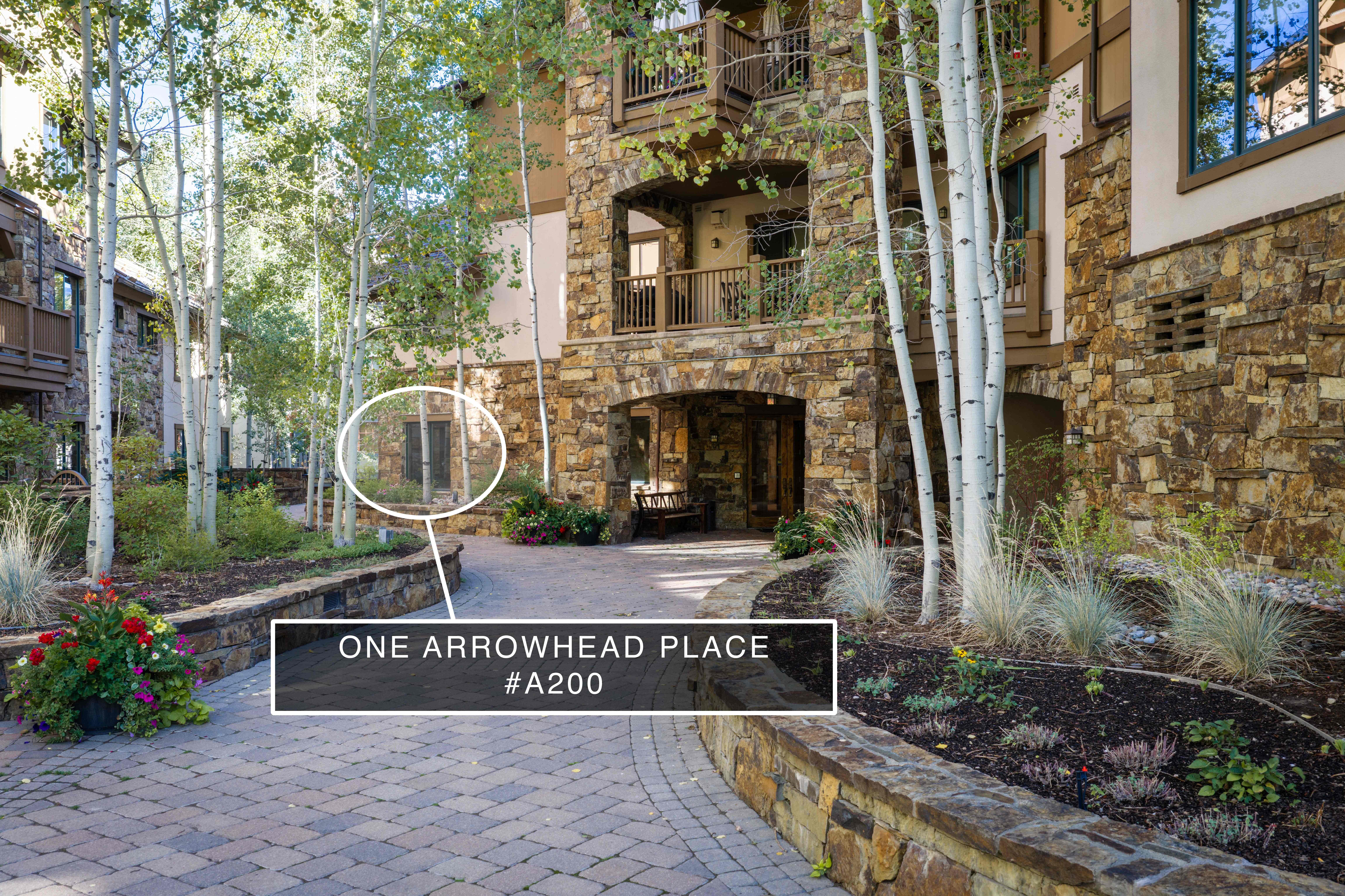 This inviting entryway features a cobblestone path lined by stone retaining walls and mature aspen trees, leading toward a rustic building entrance. The architecture incorporates natural stone cladding and timber accents, creating a warm and sophisticated mountain aesthetic. A well-maintained garden with colorful floral accents enhances the curb appeal of this welcoming residential setting.