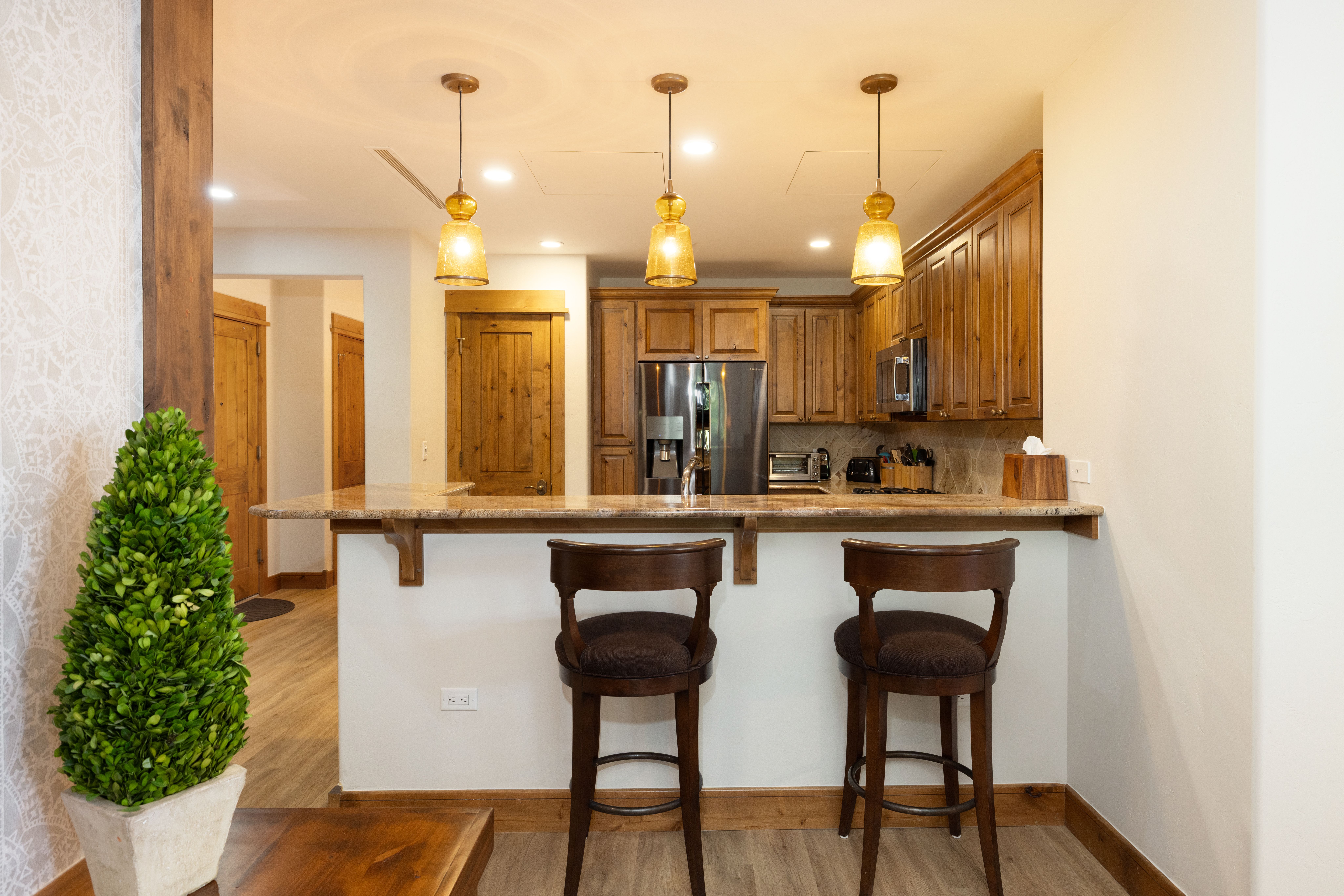 This kitchen area features a breakfast bar with two dark wood stools, complemented by warm-toned wooden cabinetry and granite countertops. Three pendant lights hang above the counter, providing a cozy and inviting atmosphere. The space transitions seamlessly into a hallway, highlighting a rustic aesthetic with wood trim and natural earth tones.