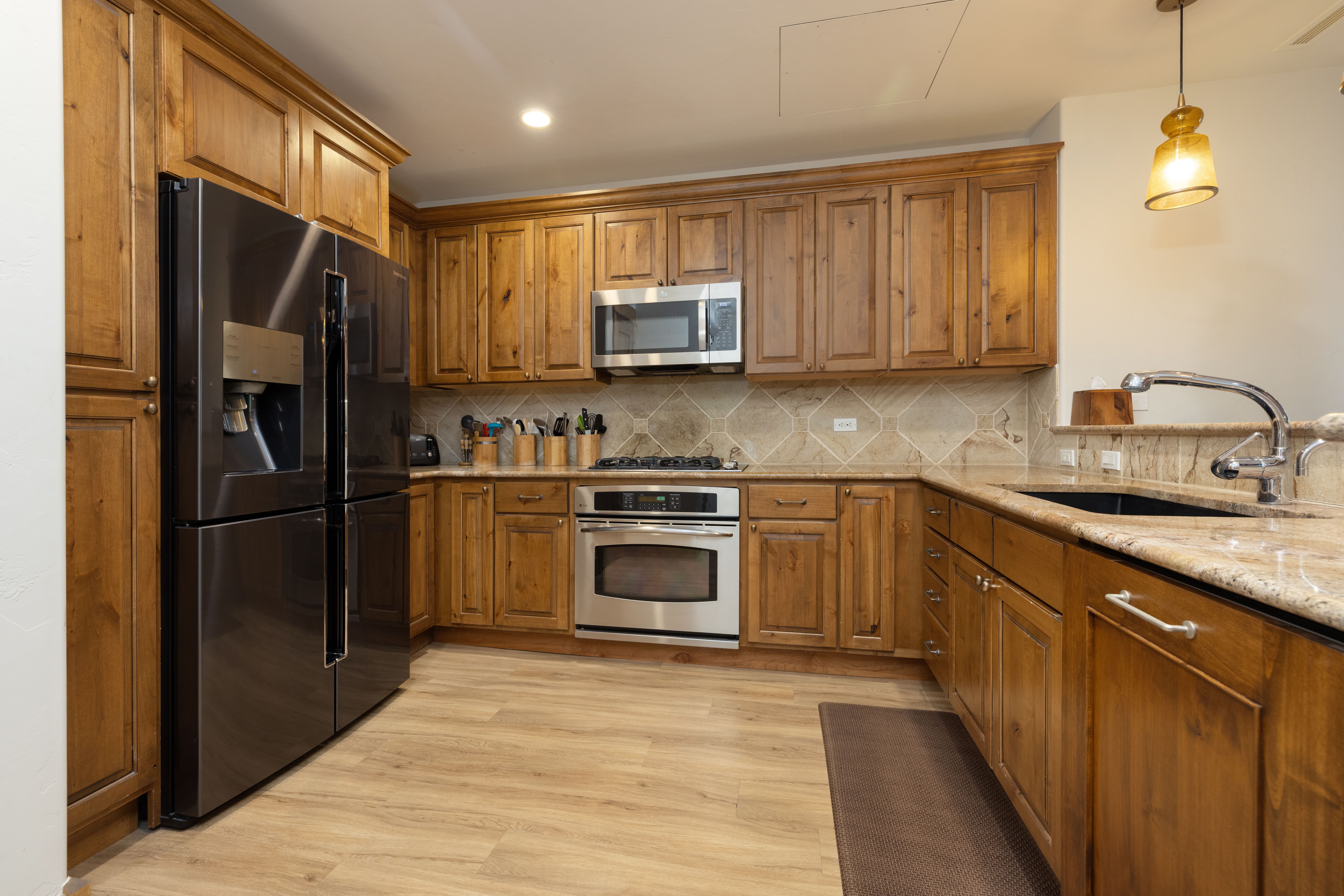 This warm and inviting kitchen features rich, natural-toned wood cabinetry paired with beige granite countertops and a coordinating tile backsplash. It is equipped with stainless steel appliances, including a microwave and oven range, alongside a sleek black refrigerator and sink. The room is completed with light wood-style flooring and a pendant light, creating a functional and cohesive workspace.