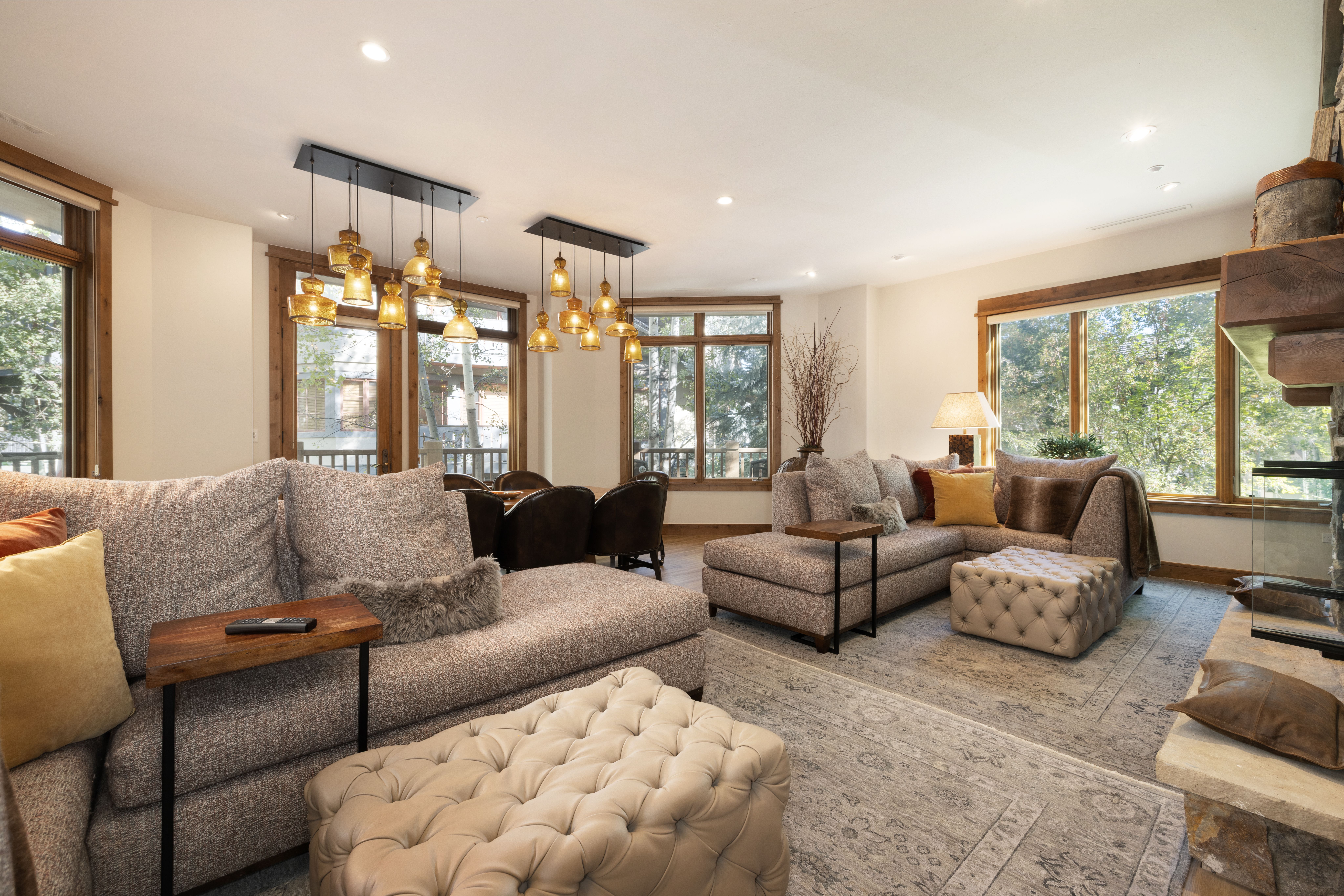This spacious and inviting living room features a cozy, open-concept design with neutral-toned sectional sofas and tufted leather ottomans. The room is highlighted by warm pendant lighting, a rustic stone fireplace, and large windows that offer scenic outdoor views. Its comfortable, mountain-inspired aesthetic makes it an ideal space for relaxation and family gatherings.