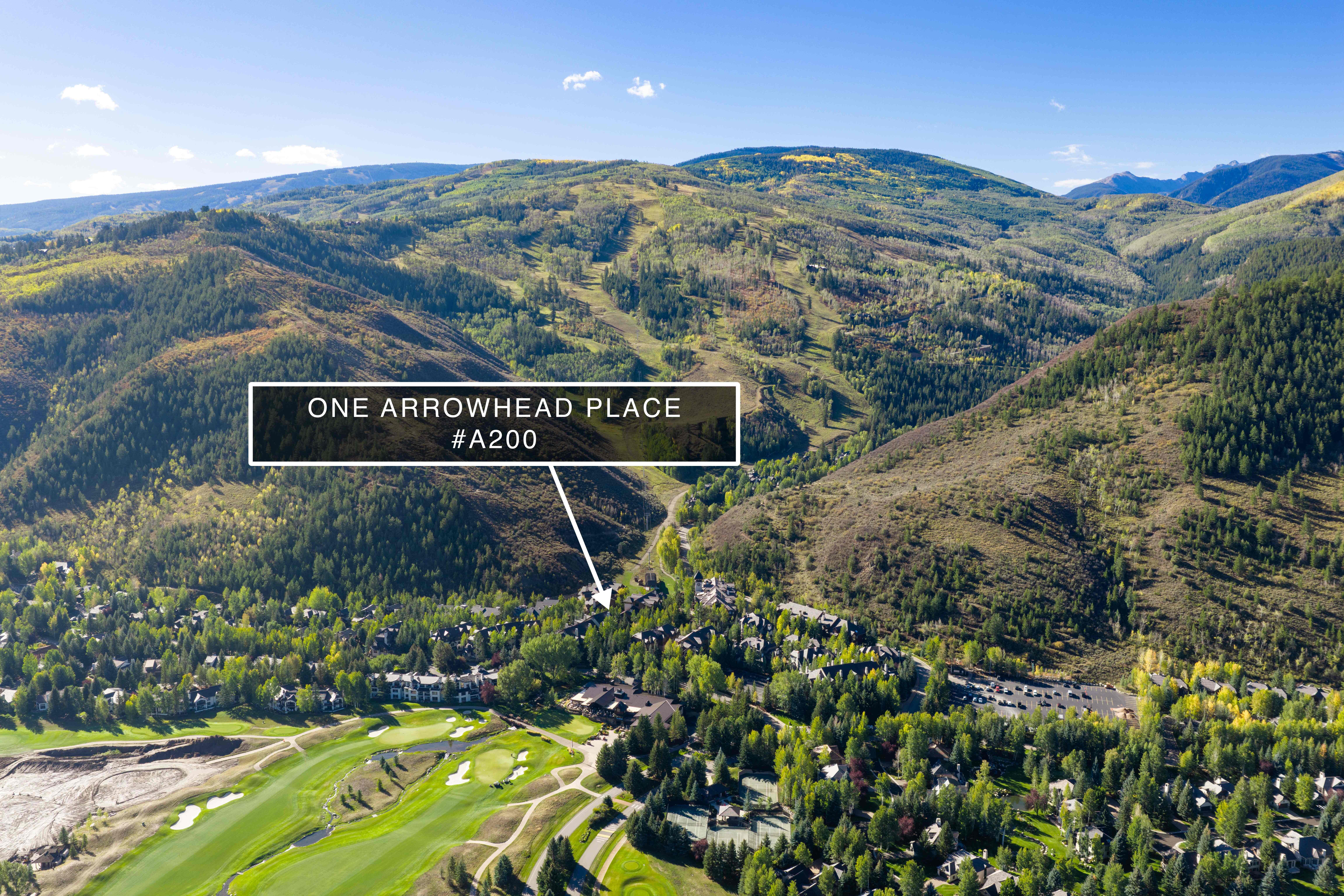 This is an expansive aerial view of a mountain resort area, highlighting a lush, forested landscape with a prominent golf course in the foreground. A graphical callout marker clearly identifies the location of 'One Arrowhead Place #A200' nestled within the scenic valley and hillside residences. The image showcases the property's prime location, offering direct access to nature and recreational facilities.