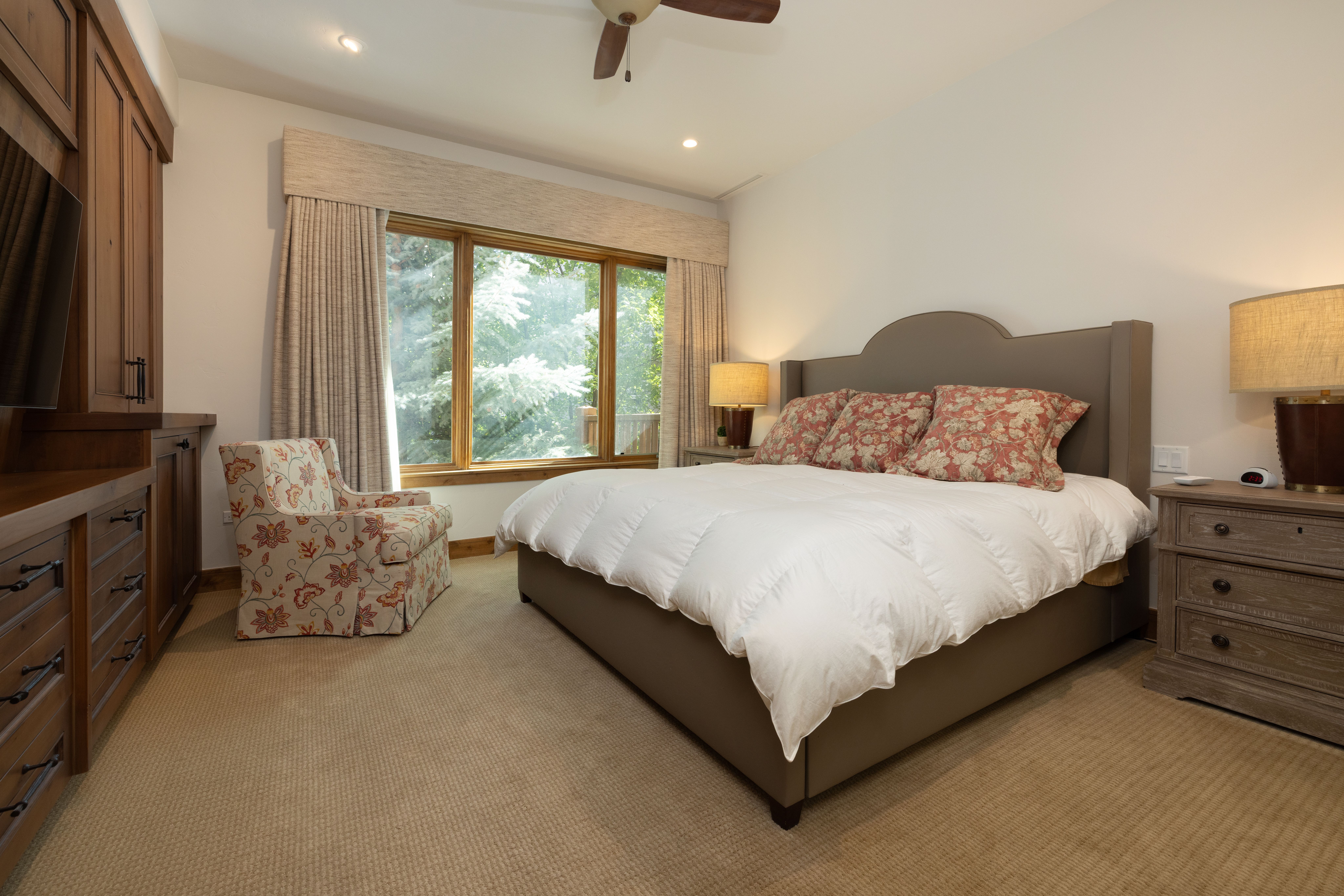This cozy primary bedroom features a large, upholstered bed with floral throw pillows, set against a backdrop of neutral walls and plush wall-to-wall carpeting. A custom wooden built-in wardrobe unit provides ample storage, while a comfortable floral armchair sits near a large window that invites natural light into the space. The room is finished with warm lighting from matching bedside lamps and a ceiling fan, creating a welcoming and comfortable retreat.