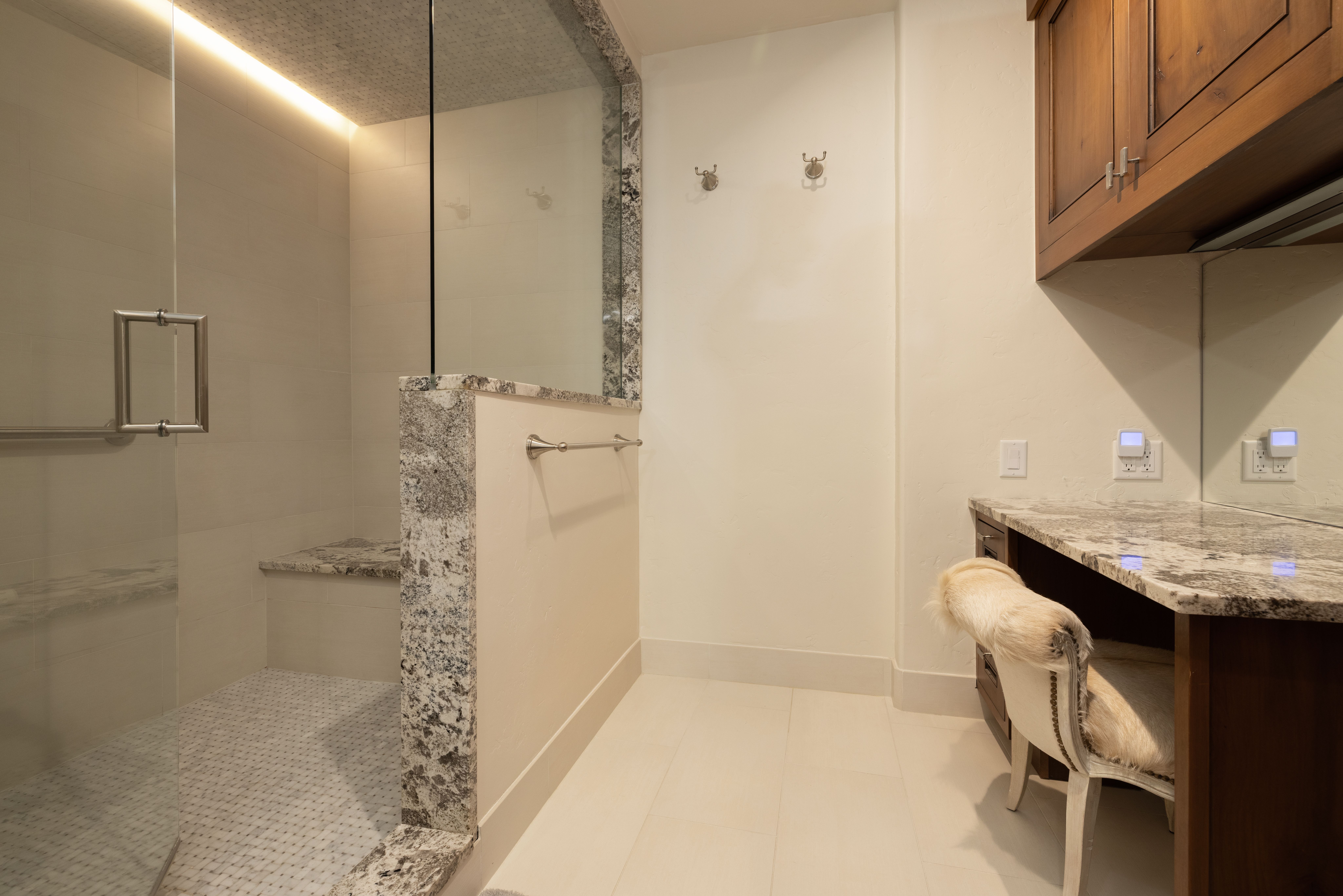 This primary bathroom features a luxurious walk-in shower with a glass enclosure, natural stone accents, and a built-in bench seat. Adjacent to the shower is a dedicated vanity area with granite countertops, dark wood cabinetry, and a unique textured chair. The combination of clean lines, high-end materials, and soft lighting creates a sophisticated and spa-like atmosphere.