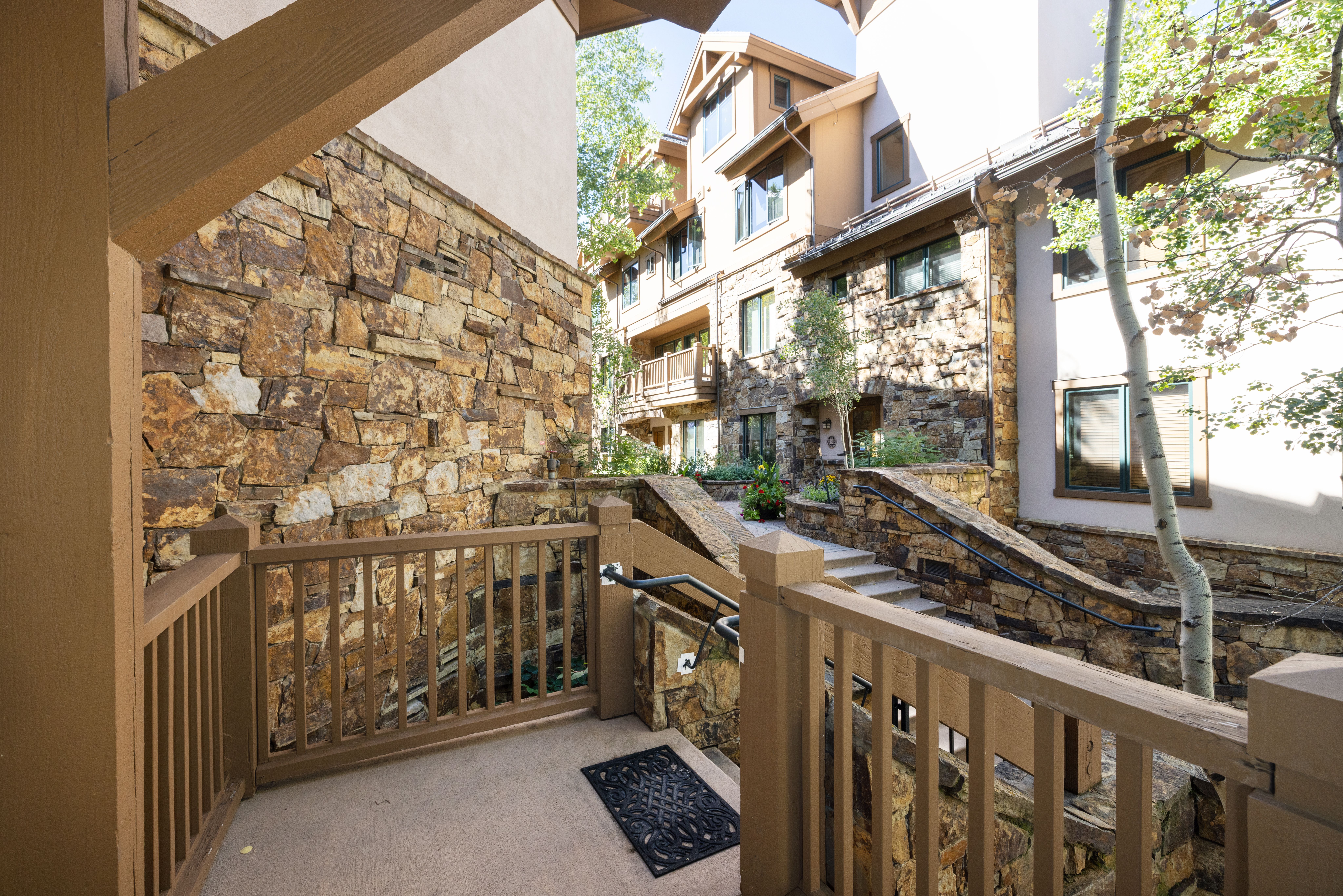 This image shows a charming entryway approach to a multi-story residential building, characterized by rustic stone masonry and classic brown wooden railings. A concrete landing with a welcome mat leads to a set of stairs that ascend towards the main building entrance, framed by lush greenery and a mature aspen tree. The architectural style conveys a cozy, mountain-lodge aesthetic, ideal for a resort or upscale dwelling.