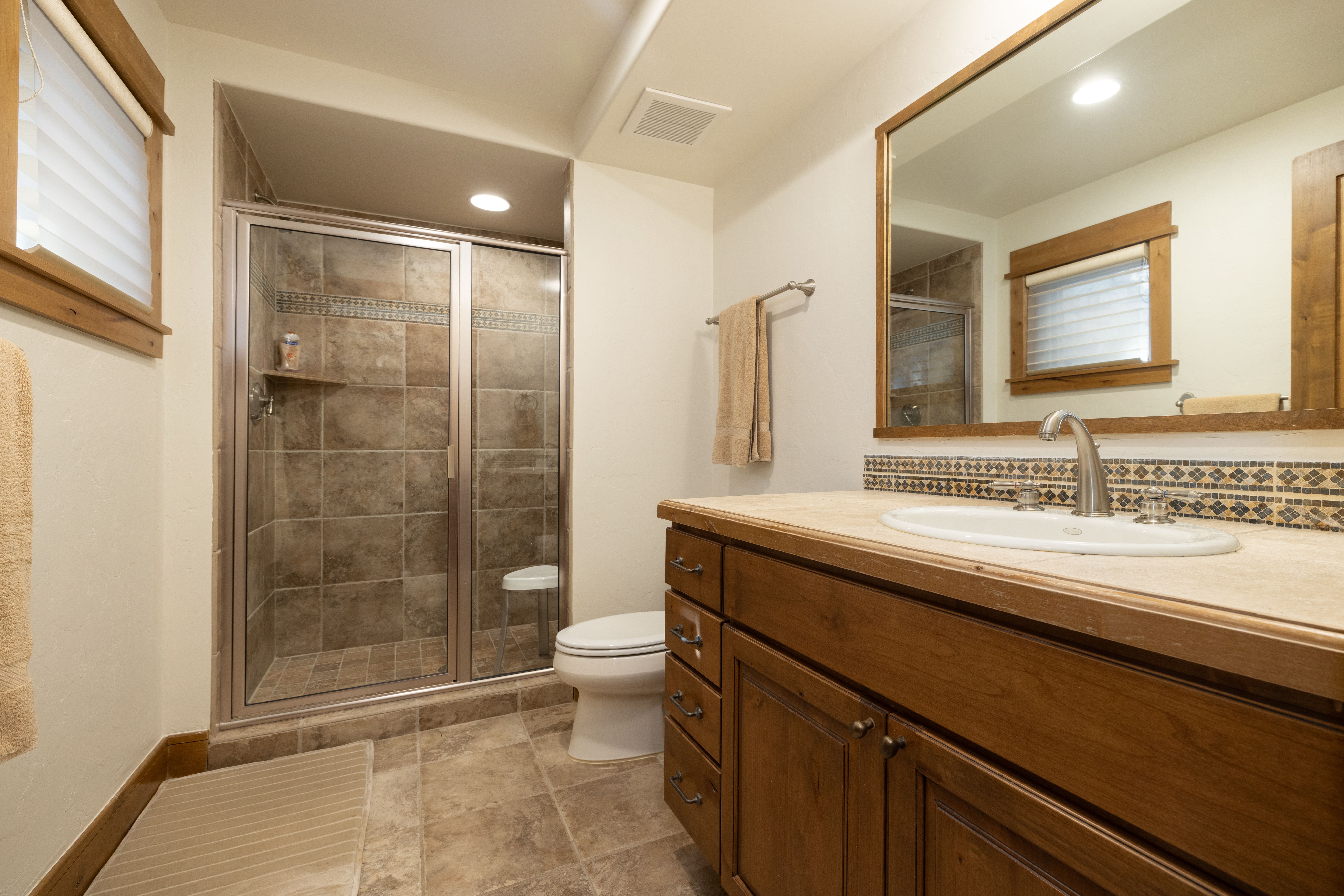 This bathroom features a rustic, desert-inspired aesthetic with warm-toned stone tile flooring and a matching tile-surround walk-in shower. A wood vanity cabinet with stone countertops provides ample storage, complemented by a large vanity mirror and natural wood trim accents. The neutral color palette and functional layout create a comfortable and inviting atmosphere.