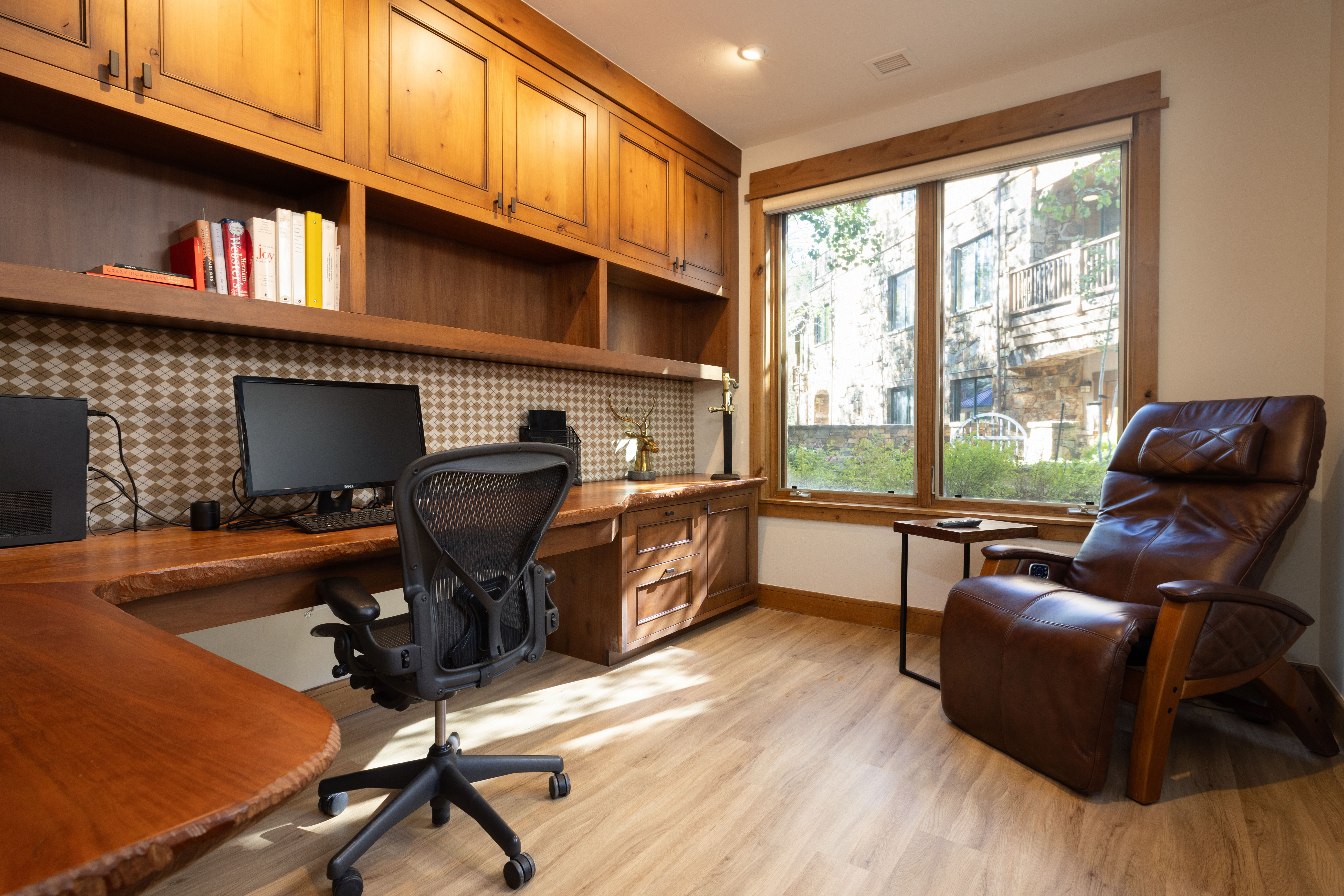 This home office features warm-toned, custom built-in wooden cabinetry and a matching live-edge wood desk, creating a sophisticated and cozy workspace. The room is equipped with an ergonomic office chair and a luxurious leather recliner by the window, which overlooks a peaceful, wooded exterior. A patterned backsplash behind the desk and light wood flooring add character and a modern, rustic charm to the study.