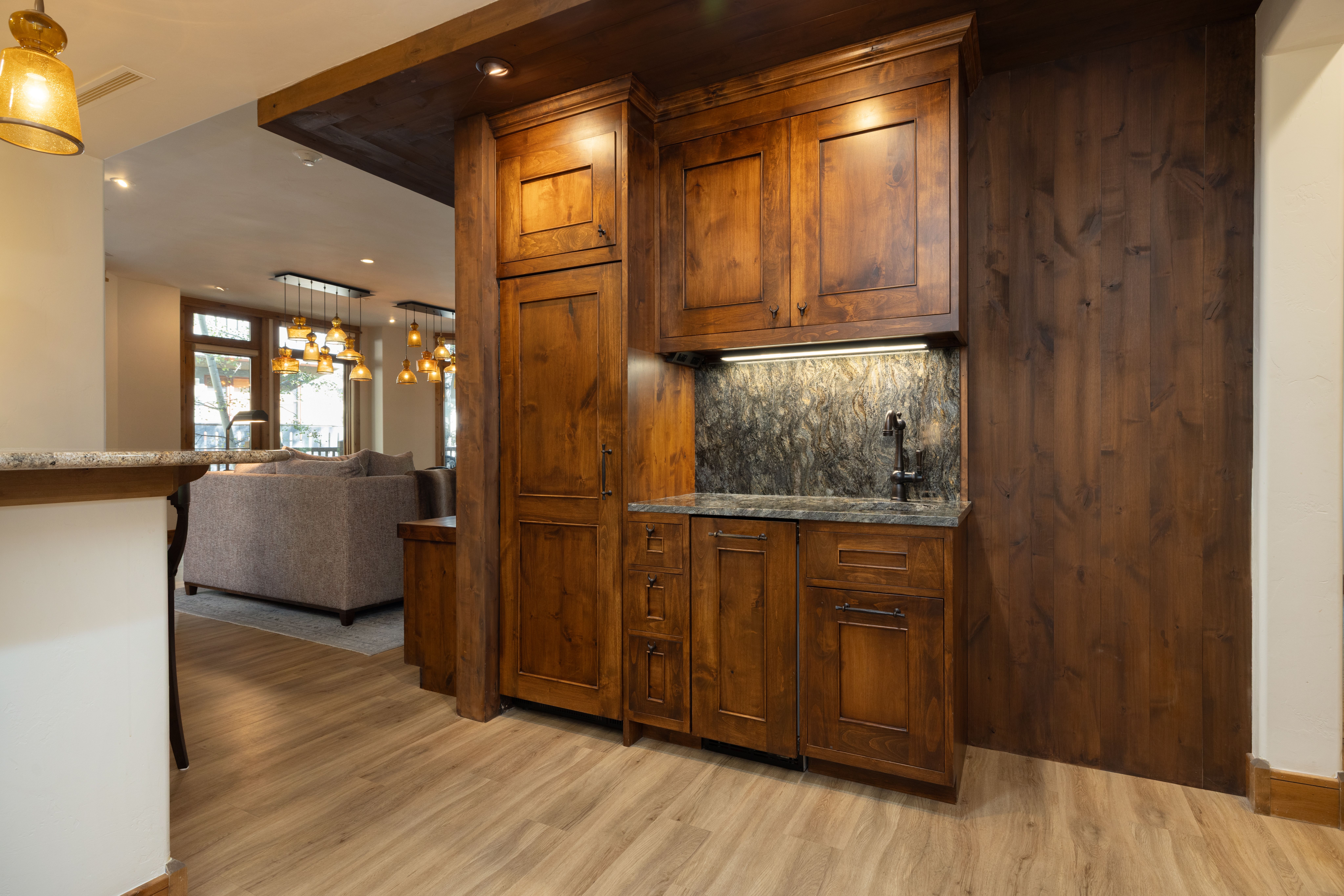 This interior space features a built-in wet bar or kitchenette area integrated with rustic, dark wood cabinetry and a rich-toned, textured stone backsplash. The area includes a small sink with a dark faucet, ample drawer and cabinet storage, and warm under-cabinet lighting. Adjacent to a living room, this refined wood-paneled space adds a cozy and sophisticated element to the home's open floor plan.