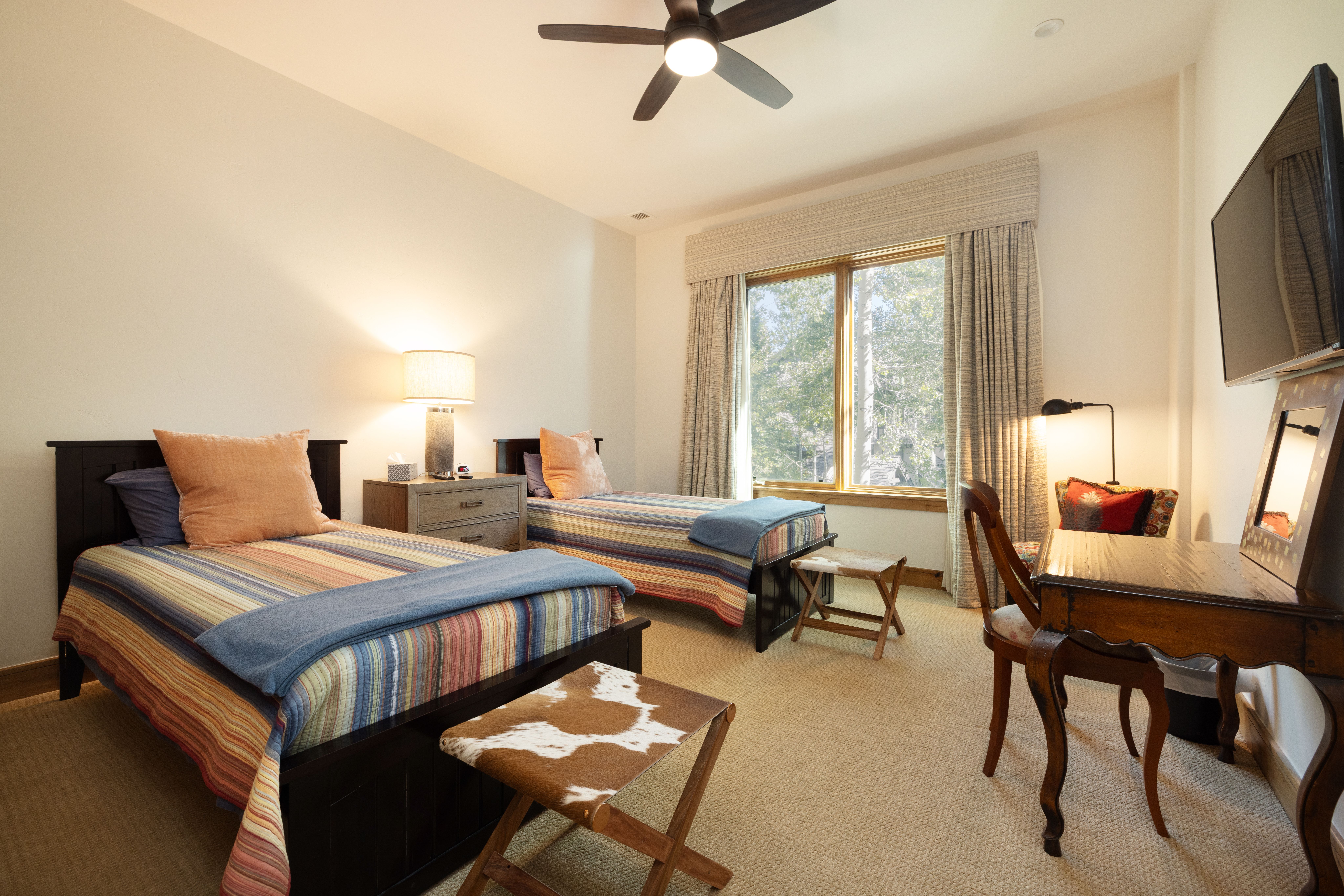 This guest bedroom features two twin beds with coordinated striped bedding and warm, rustic accents like the cowhide footstools. The room is well-appointed with a dedicated desk area, professional window treatments, and a ceiling fan, creating a comfortable and inviting space. The neutral color palette and functional layout make it an ideal retreat for guests.
