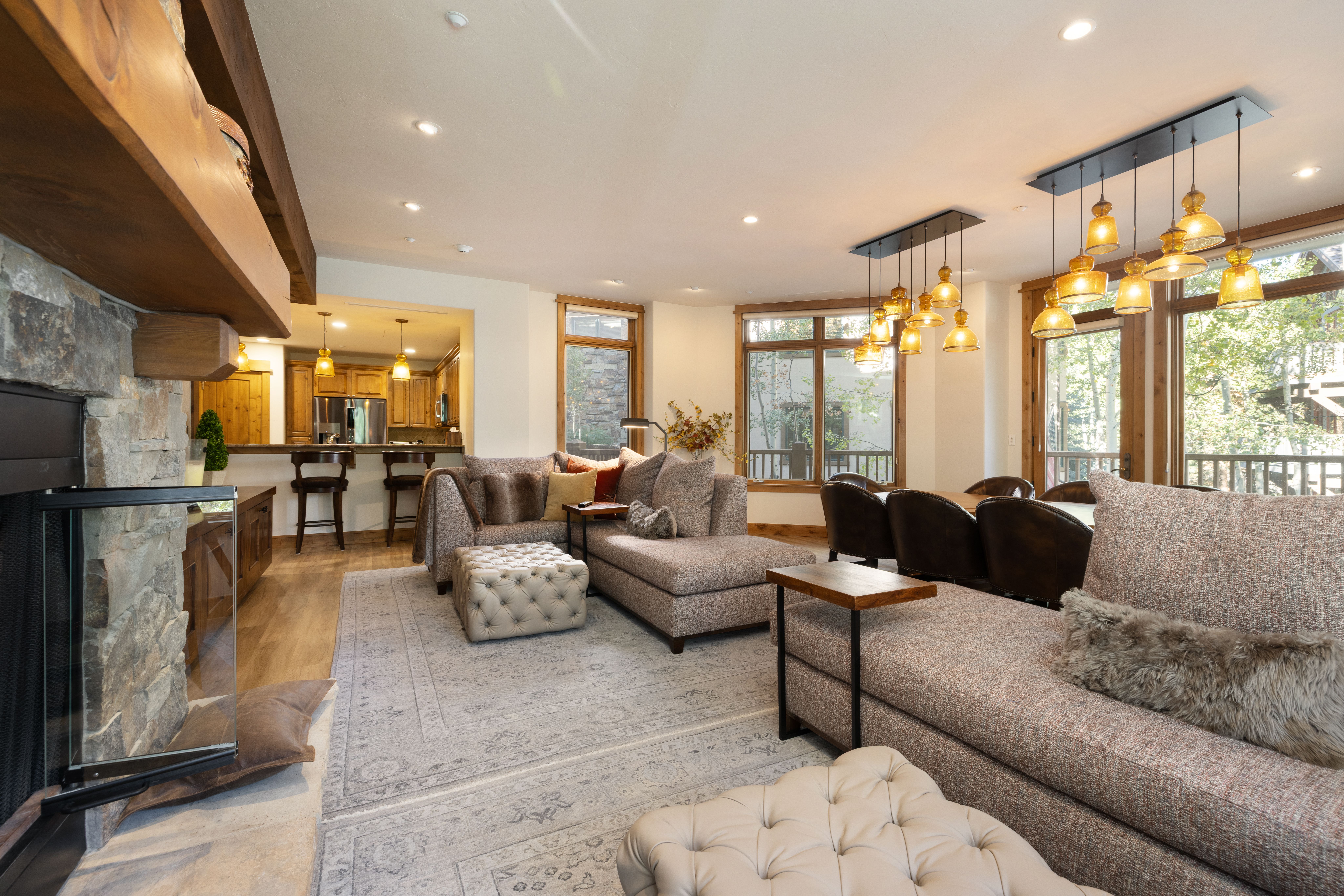 This spacious and inviting living area features an open floor plan that seamlessly integrates with a dining area and kitchen. Highlights include a stunning floor-to-ceiling stone fireplace, plush neutral-toned sectionals, and an elegant patterned area rug. The space is filled with natural light from large windows and is accented by sophisticated pendant lighting, creating a warm, mountain-modern atmosphere.