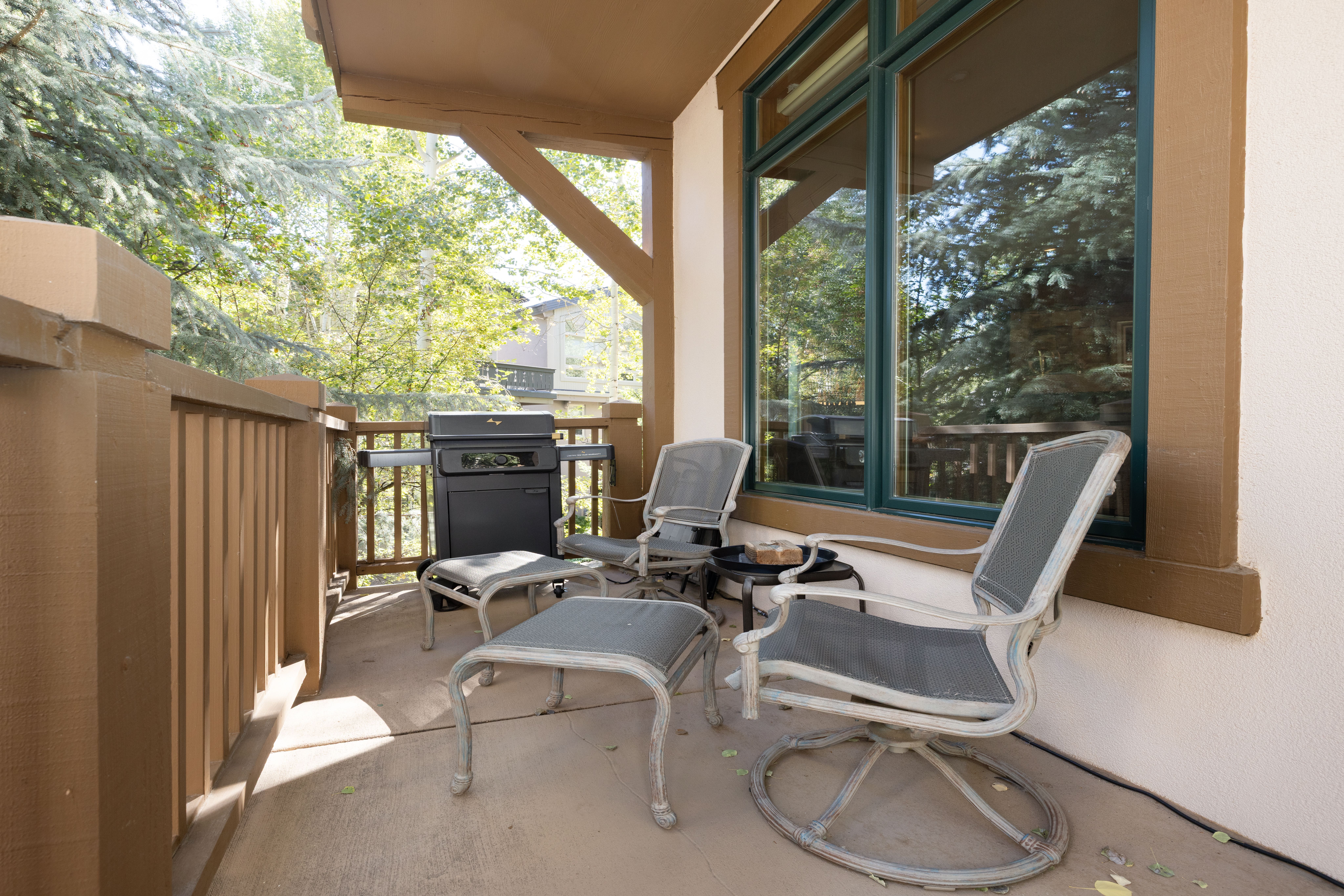 This covered balcony offers a peaceful outdoor seating area with two comfortable chairs and matching ottomans. A small side table holds a wooden decor piece, and a grill is tucked away in the corner for convenient outdoor cooking. Large windows provide a view of the surrounding trees, creating a quiet and relaxing space perfect for enjoying the outdoors.