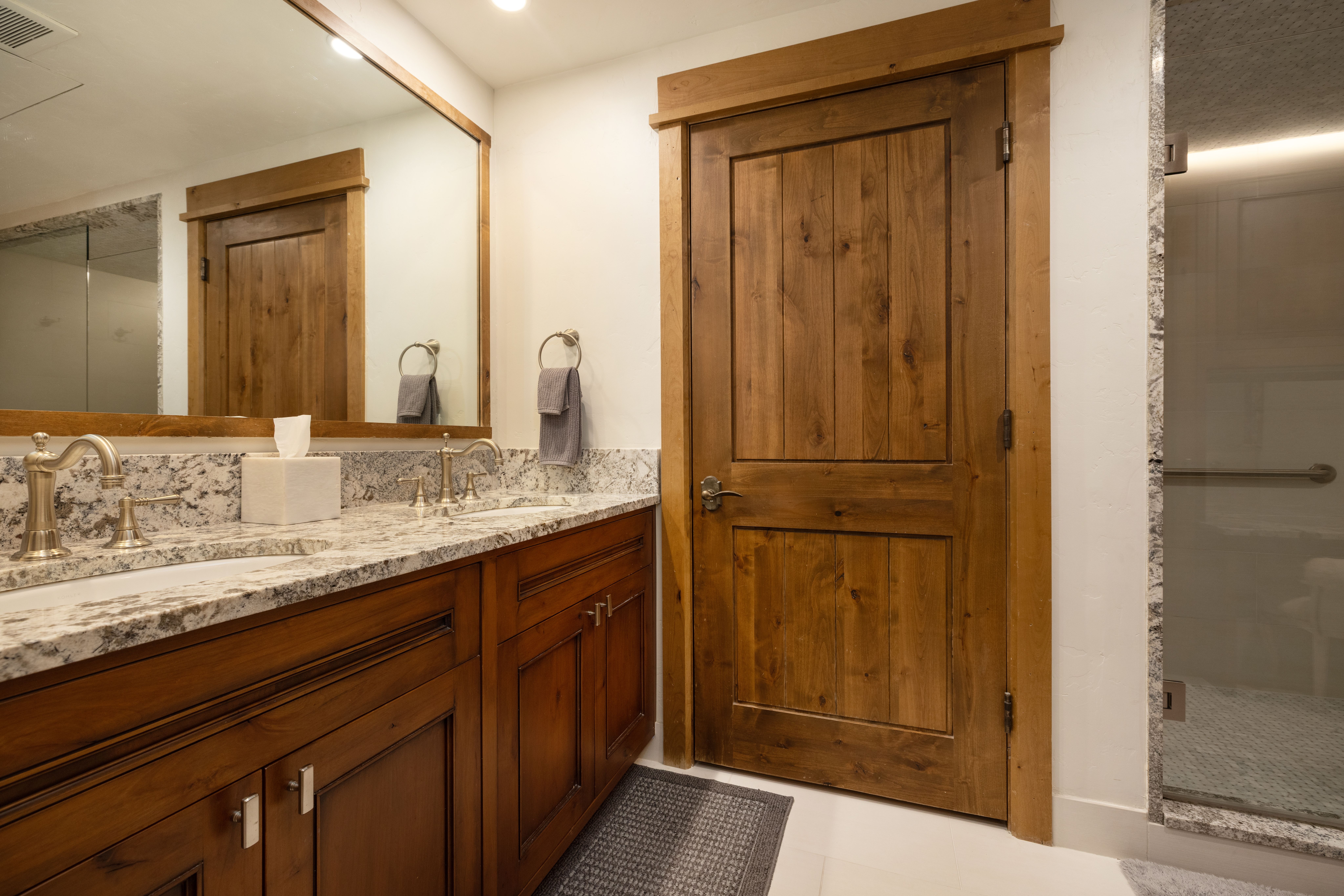 This primary bathroom features a warm, rustic aesthetic with a dual-sink vanity topped with elegant granite countertops and traditional metal fixtures. The space is anchored by a wooden door with a craftsman-style frame and a large, full-width mirror that enhances the room's brightness. A glass-enclosed shower is visible on the right, providing a cohesive and functional layout suitable for a high-end residence.