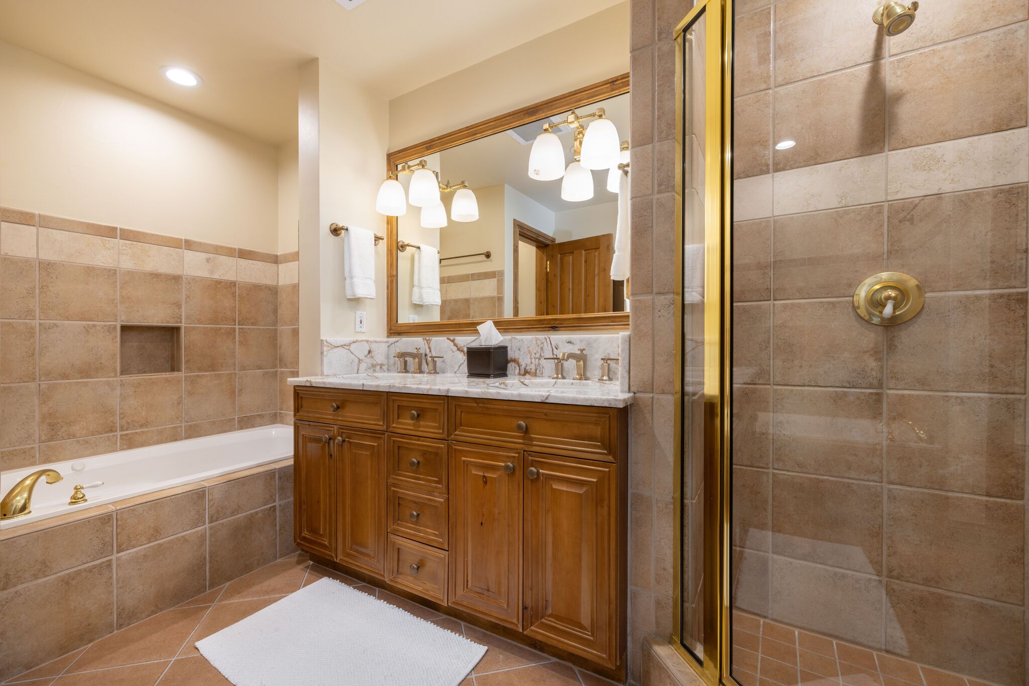 This primary bathroom features a warm, rustic-inspired design with rich wood cabinetry, a decorative marble backsplash, and large-format tan wall tiles. The space includes a built-in soaking tub, a double-sink vanity with gold-toned hardware, and a separate glass-enclosed shower, creating a comfortable and coordinated traditional aesthetic.
