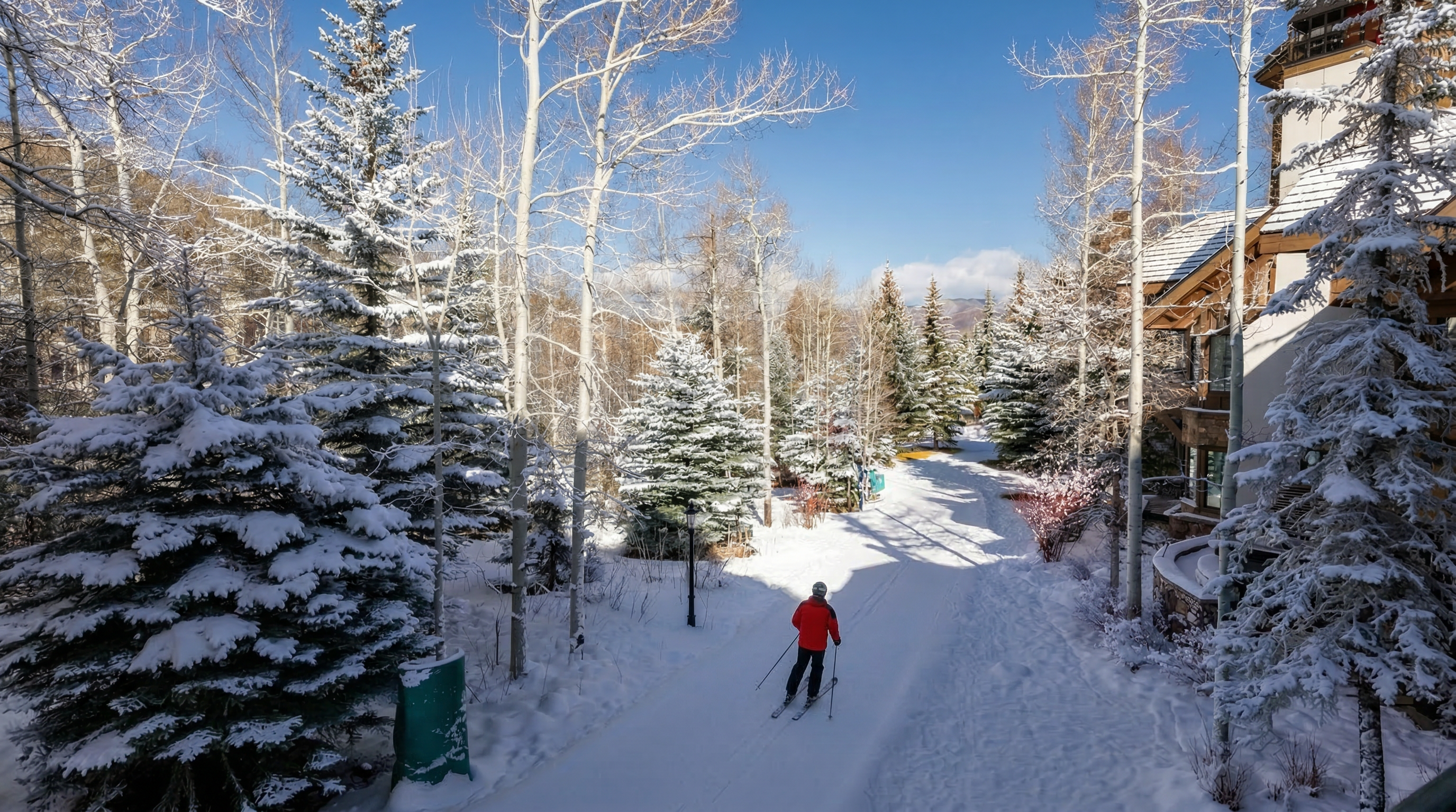 This scenic image captures a snowy pass at a high-end ski resort, featuring a groomed path winding between frost-covered pine trees and elegant timber-framed architecture. A skier in a bright red jacket is seen navigating the serene landscape, highlighting the convenient ski-in/ski-out access often prized in mountain real estate. The clear blue sky and pristine snowy environment create a peaceful and luxurious winter atmosphere.
