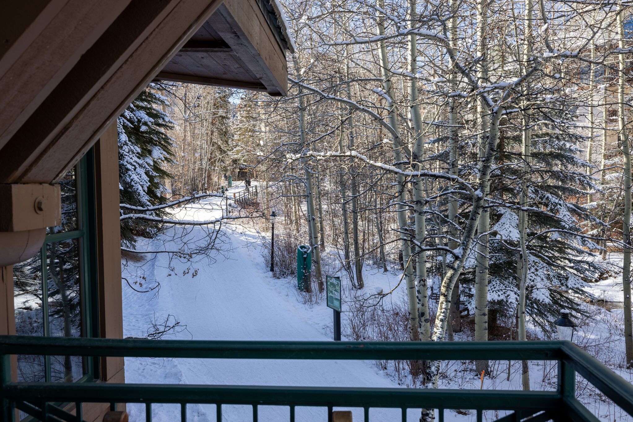 This view, captured from an elevated balcony, overlooks a serene, snow-covered pathway winding through a wooded landscape. The scene features a mix of evergreen trees and bare aspen trunks, offering a peaceful winter setting typical of a mountain resort. A portion of the building's covered balcony framing remains visible in the foreground, providing context to this scenic outdoor vantage point.