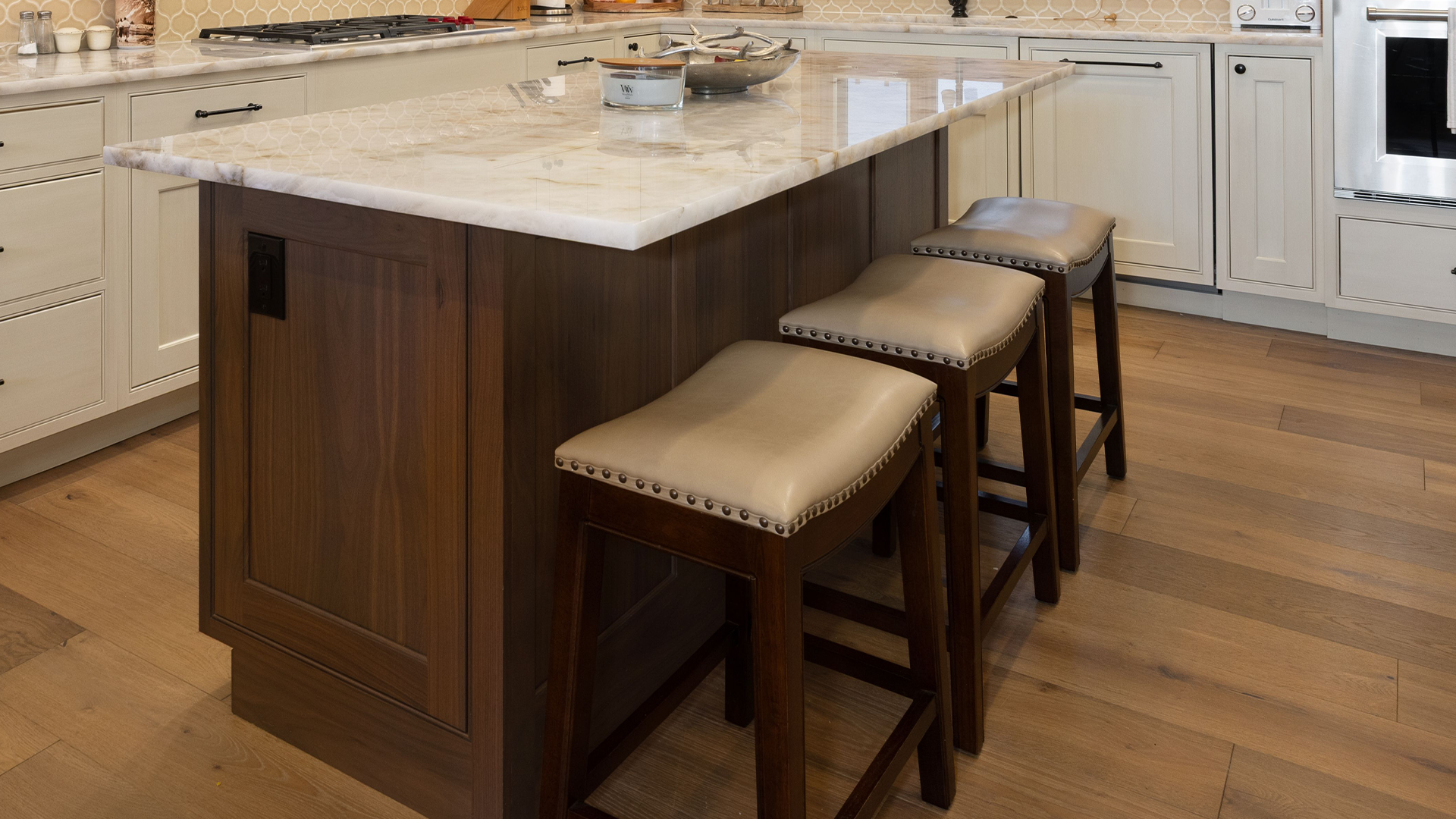 A sophisticated kitchen island serves as a central focal point, featuring rich, dark wood cabinetry contrasted by a luxurious light-veined marble countertop. The island is complemented by three elegant stools with nailhead trim, while the surrounding kitchen area showcases seamless white cabinetry and high-end stainless steel appliances. The warm wood flooring and tasteful backsplash create a refined and cohesive aesthetic suitable for a modern luxury home.