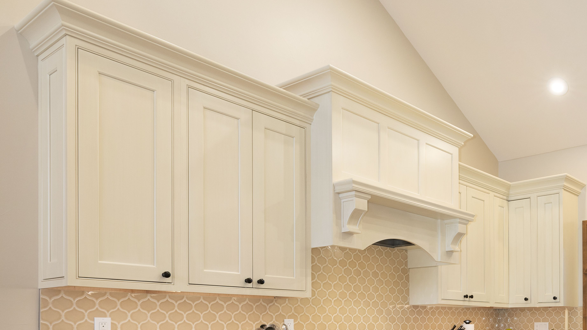 This kitchen features elegant, off-white shaker-style cabinetry with detailed crown molding that reaches up to the vaulted ceiling. A custom-built range hood with decorative corbels serves as a focal point above a decorative, scalloped mosaic tile backsplash. The design offers a bright and timeless aesthetic, characteristic of a high-quality residential kitchen layout.