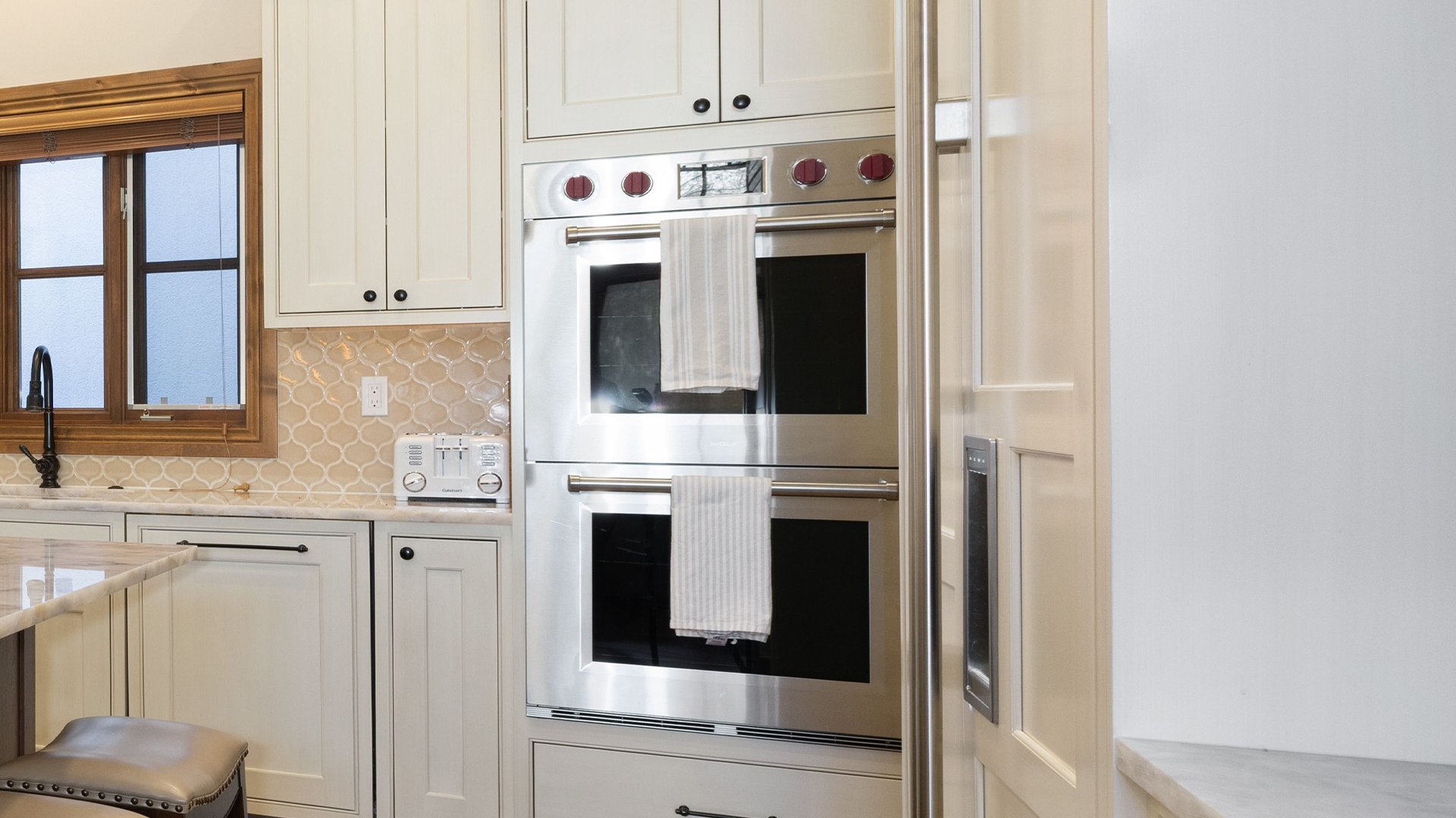 This kitchen features a clean, classic design with elegant off-white cabinetry and a high-end stainless steel double wall oven. A decorative scalloped-tile backsplash adds texture and sophistication, while a window with wooden casing brings natural light and warmth to the space. The overall aesthetic is polished, functional, and well-maintained.