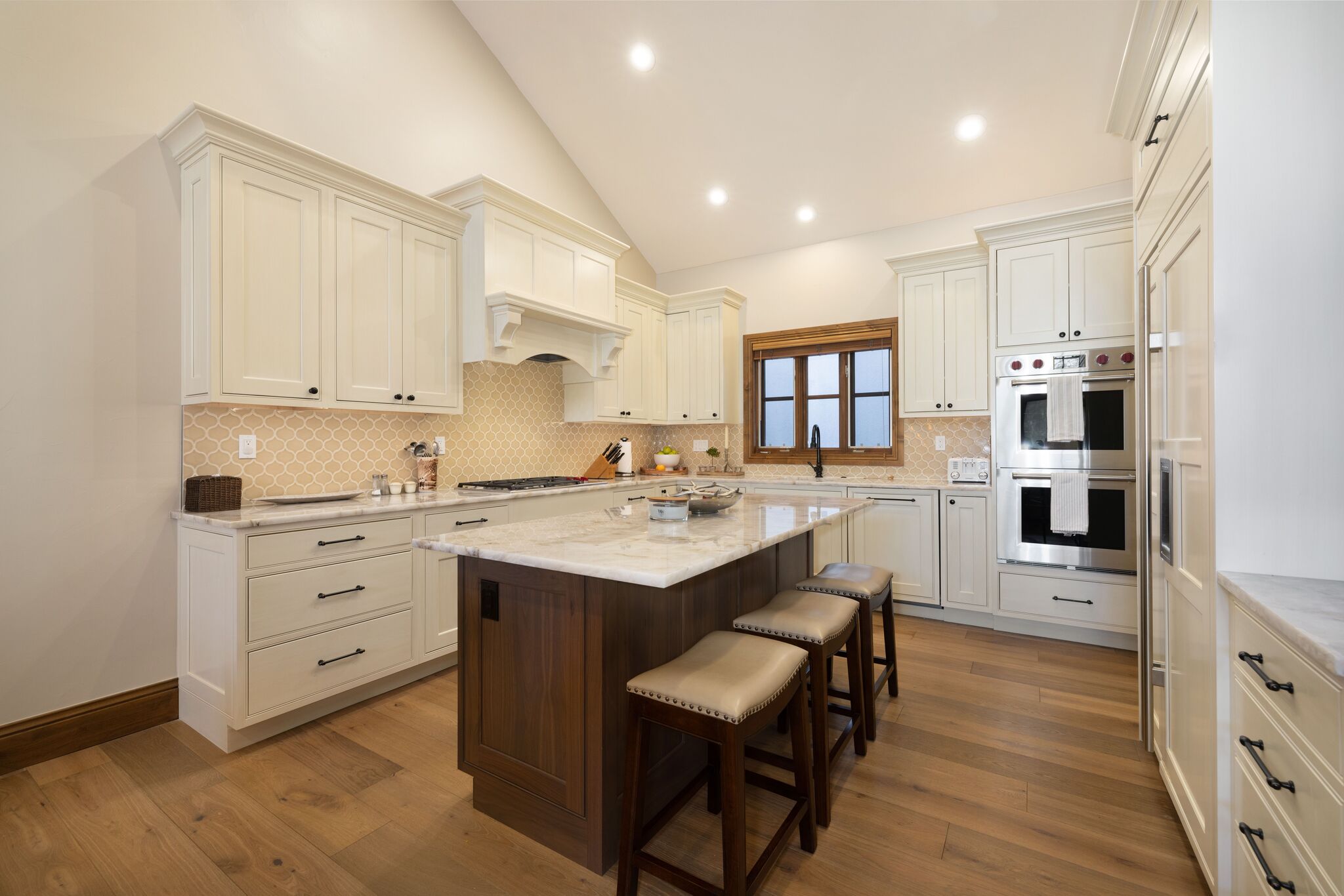 This spacious, high-end kitchen features creamy off-white cabinetry, gleaming marble countertops, and a rich, dark wood kitchen island with bar stool seating. The area is illuminated by recessed lighting and a classic backsplash, creating an elegant and inviting atmosphere. A built-in double oven and ample storage make this a functional and stylish space perfect for cooking and entertaining.