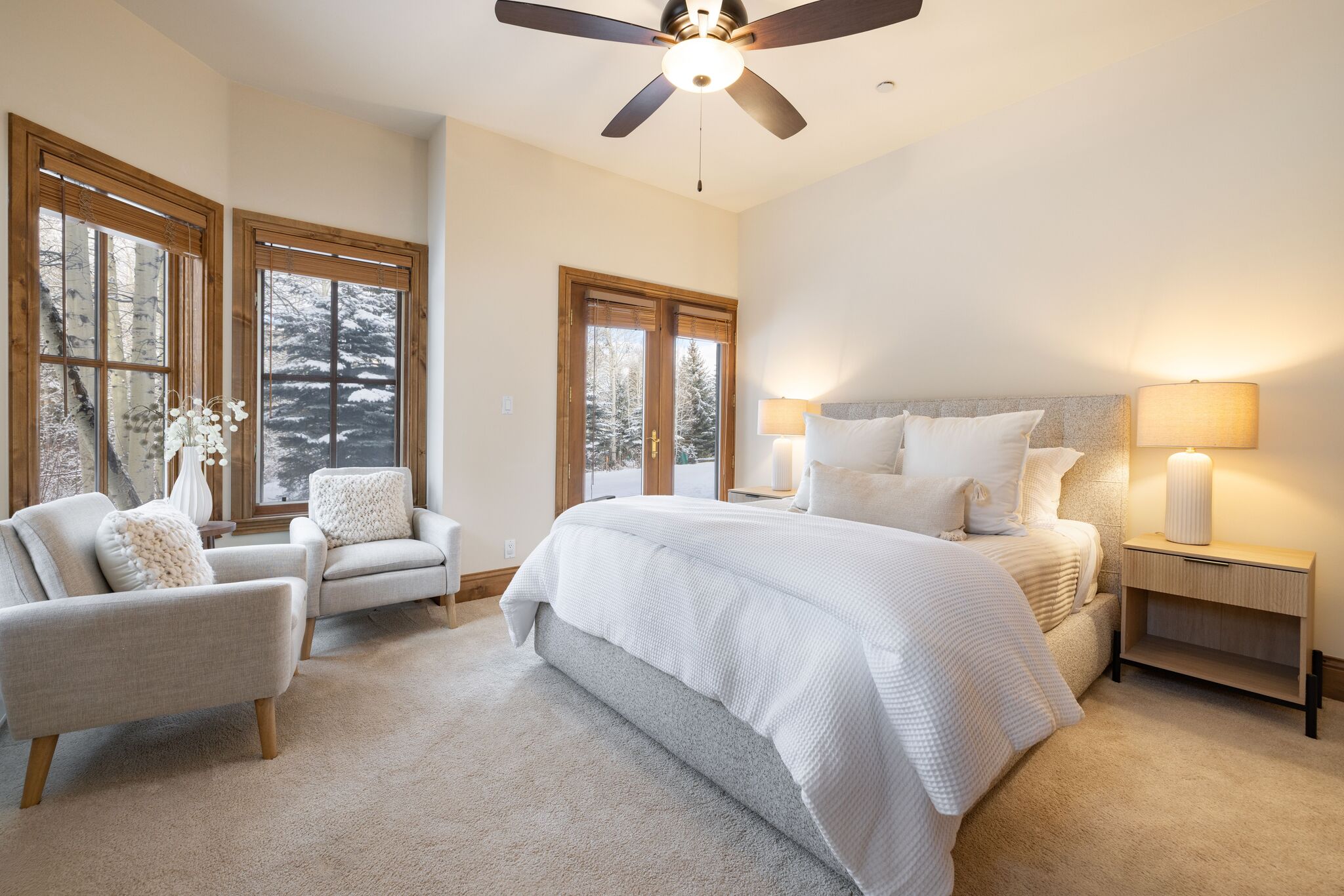 This bright and inviting primary bedroom features a plush, textured queen bed with neutral-toned bedding and two matching modern nightstands. A cozy seating arrangement with two armchairs near the windows adds a comfortable touch, while large windows provide ample natural light and views of a snow-covered landscape. The room is completed by a ceiling fan and elegant bedside lighting, creating a warm and serene retreat.