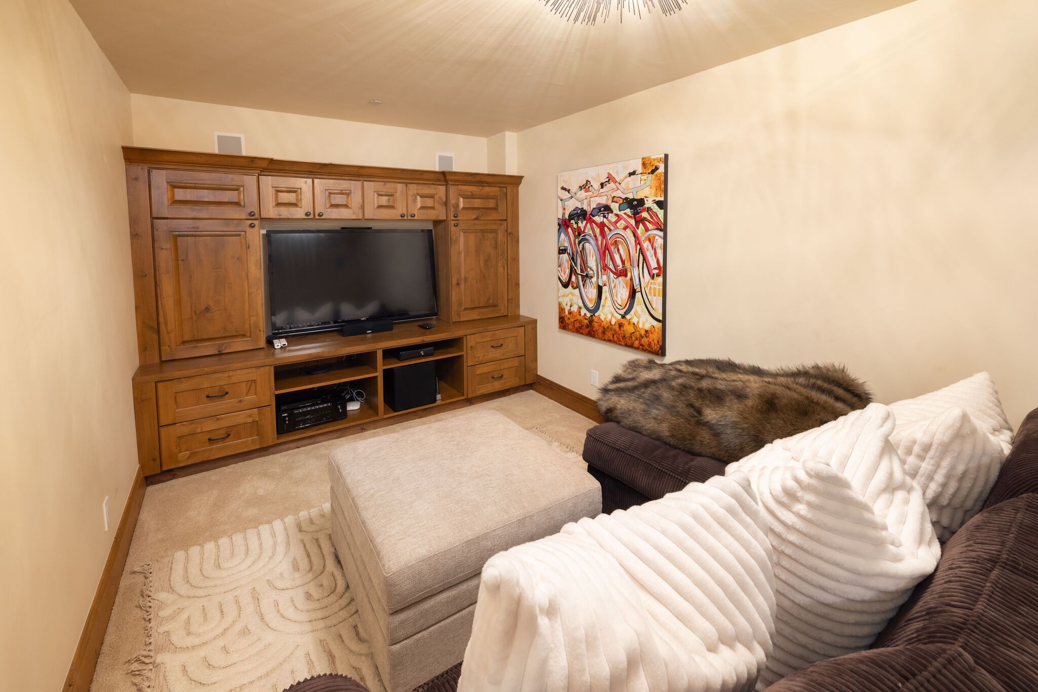 This cozy living area features a large, custom-built wooden entertainment center that houses a flat-screen television and media storage. The room is comfortably furnished with a plush dark sofa topped with faux fur throws, a matching light-colored ottoman, and a textured area rug, creating an inviting space for relaxation. A colorful, bicycle-themed canvas art piece adds a touch of character to the neutral beige walls.