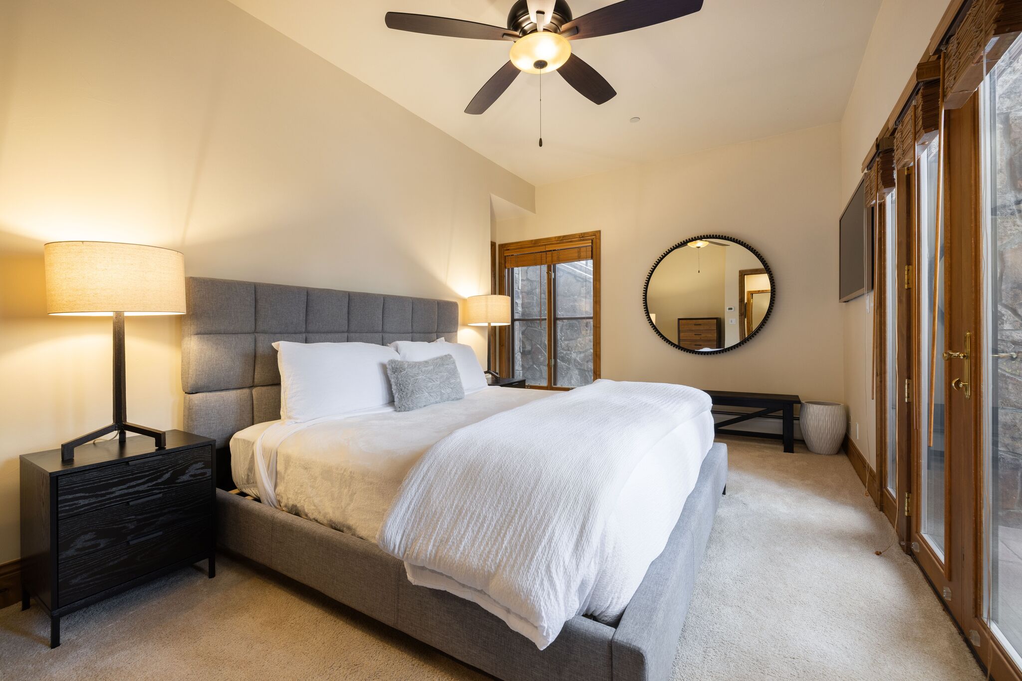 This primary bedroom features a modern gray upholstered bed with crisp white bedding, anchored by a sleek black nightstand and matching table lamp. A large circular mirror on the wall adds a contemporary touch, while natural light streams in from the expansive sliding glass doors that lead to an outdoor space. The light carpeting and neutral wall color create a calming, refined atmosphere suitable for a comfortable living space.