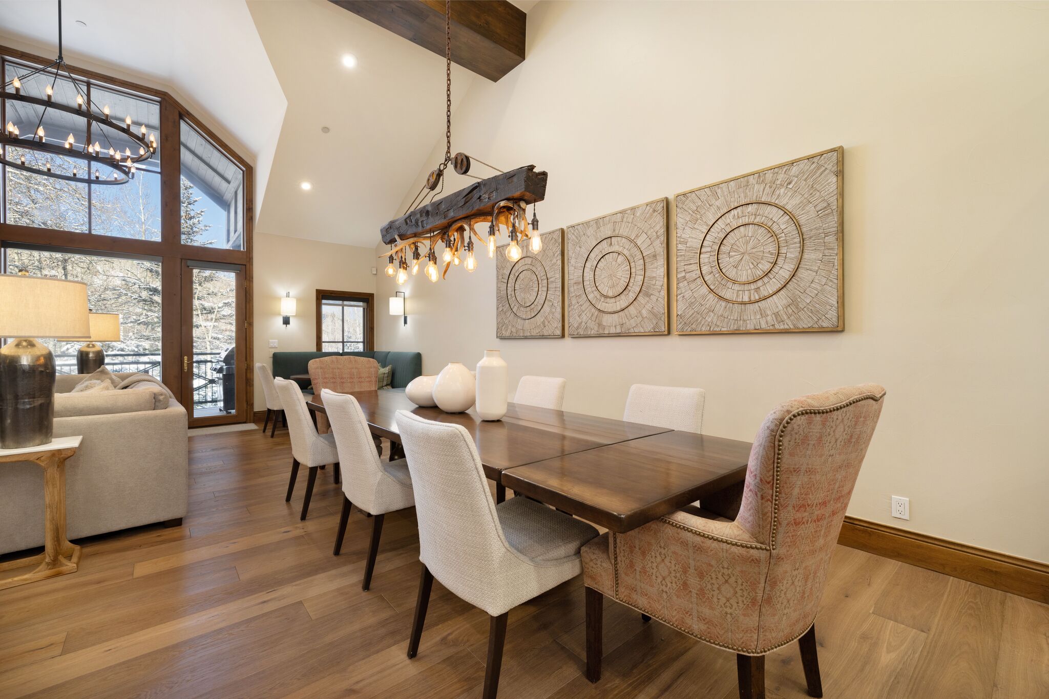 This elegant dining room features a long, dark wood table surrounded by a mix of light-upholstered side chairs and patterned armchairs, creating a balanced and inviting space. The room is anchored by a unique, rustic chandelier made from a reclaimed timber beam and antler-inspired fixtures. Large wall art pieces with circular, textured motifs provide a sophisticated focal point, while the open floor plan flows seamlessly into a living area with a view of a snowy landscape.
