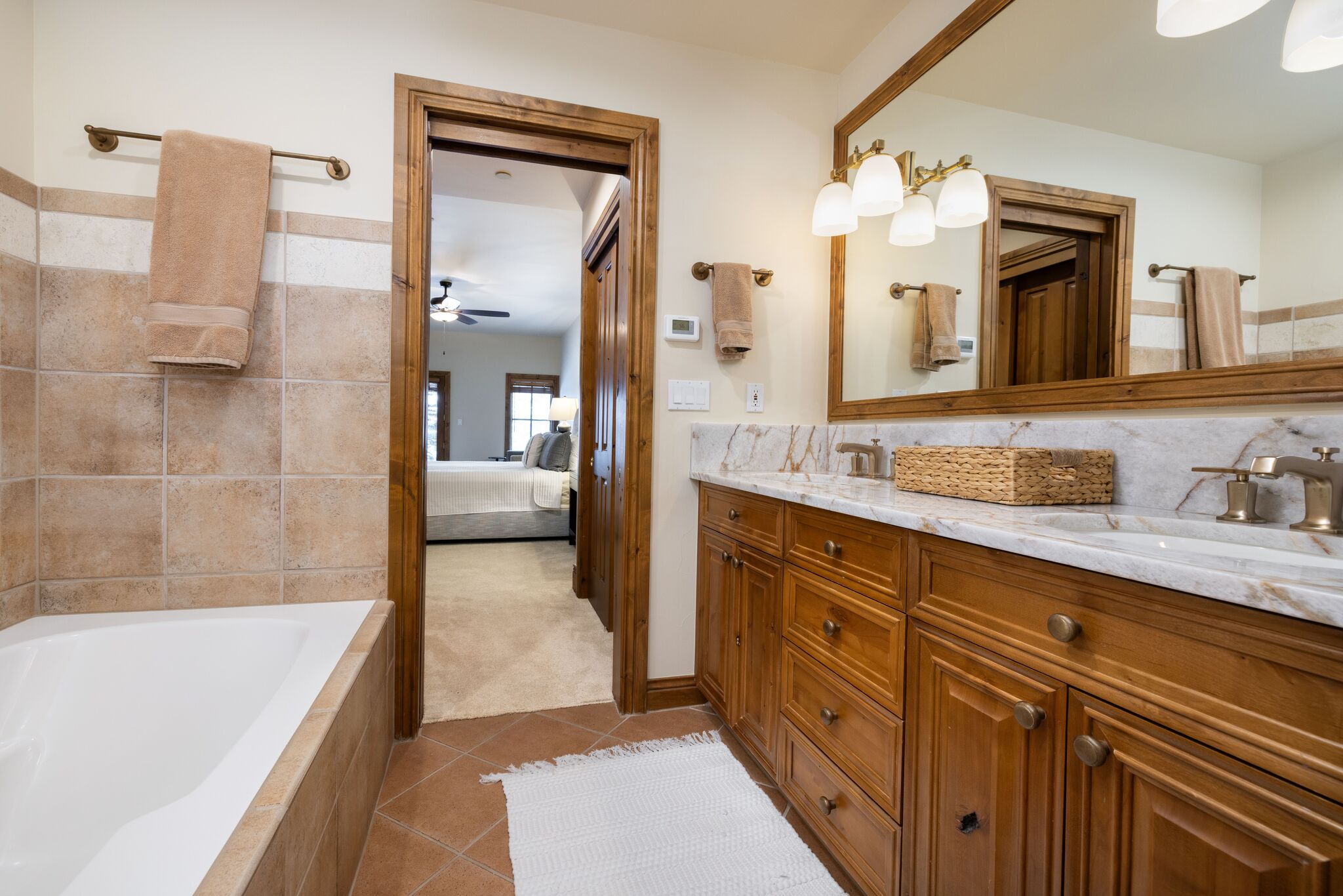 This primary bathroom features a warm, rustic aesthetic with natural wood cabinetry, tan tile work, and a marble countertop with dual sinks. The space includes a soaking tub, elegant brass-toned fixtures, and a clear view into an adjacent bedroom through an open doorway, creating a cohesive and inviting layout.