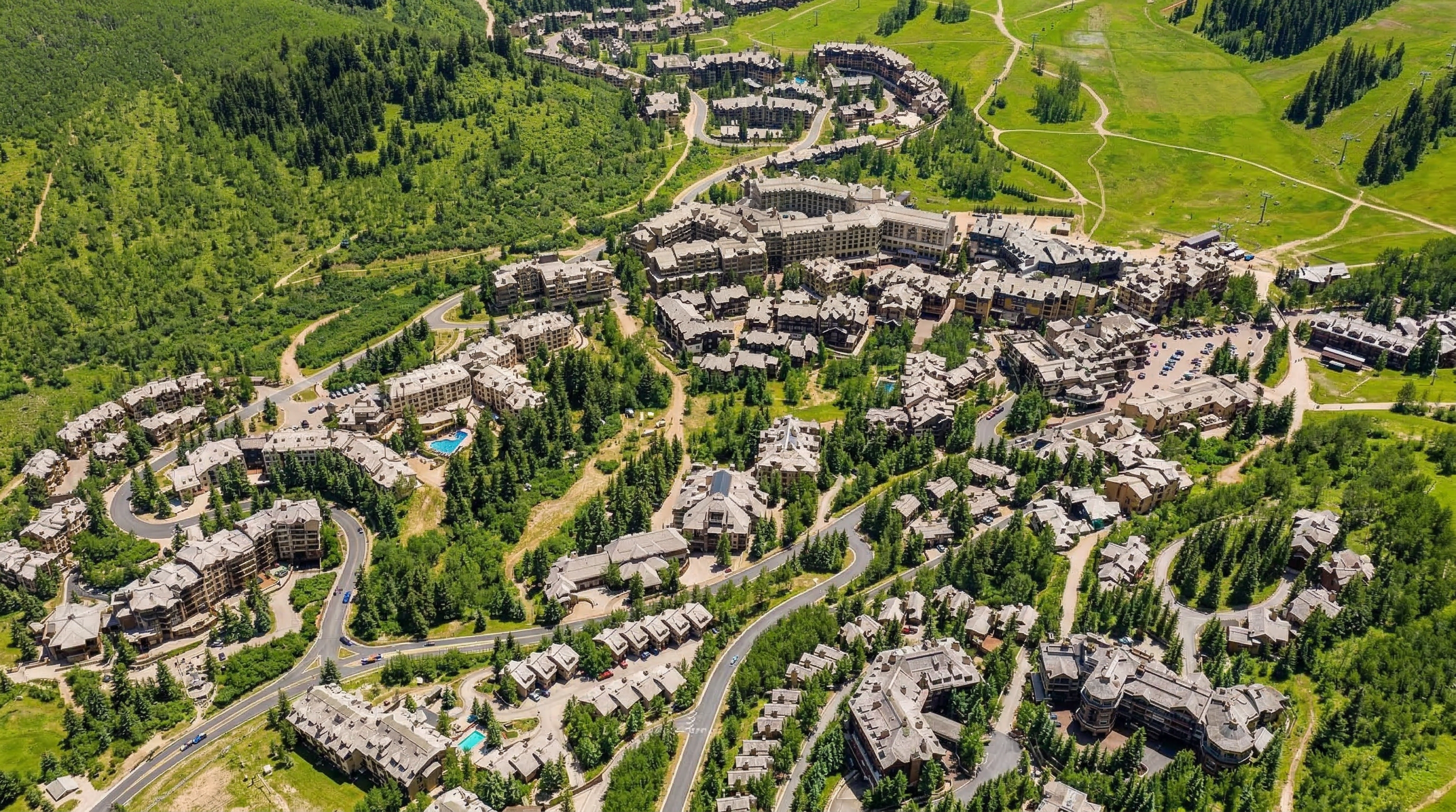 This stunning aerial view highlights a sprawling mountain-style residential resort nestled deep within a lush, forested landscape. The community features a dense collection of large lodges and townhomes with classic wood and stone architecture, seamlessly integrated into the natural scenery. Winding roads connect the various clusters of buildings, creating an inviting and picturesque high-altitude retreat.