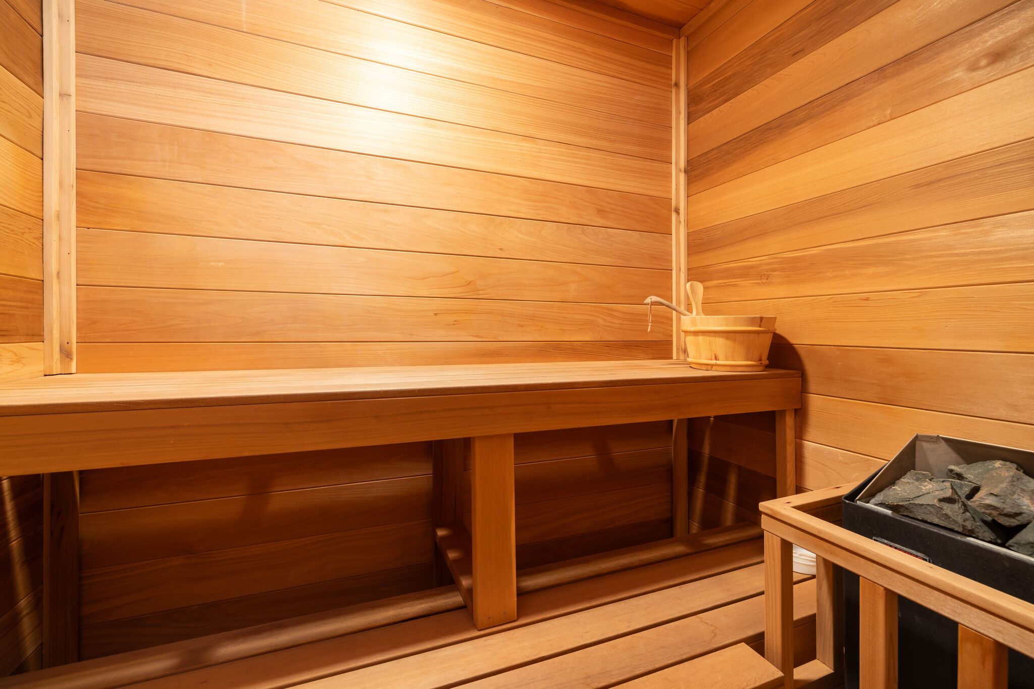 This image features a traditional cedar sauna, showcasing high-quality wood paneling throughout the space. The room includes a built-in wooden bench and a wood-fired heater with stone, creating an authentic and relaxing spa-like environment. It is a well-maintained and classic feature, indicative of luxury home amenities.