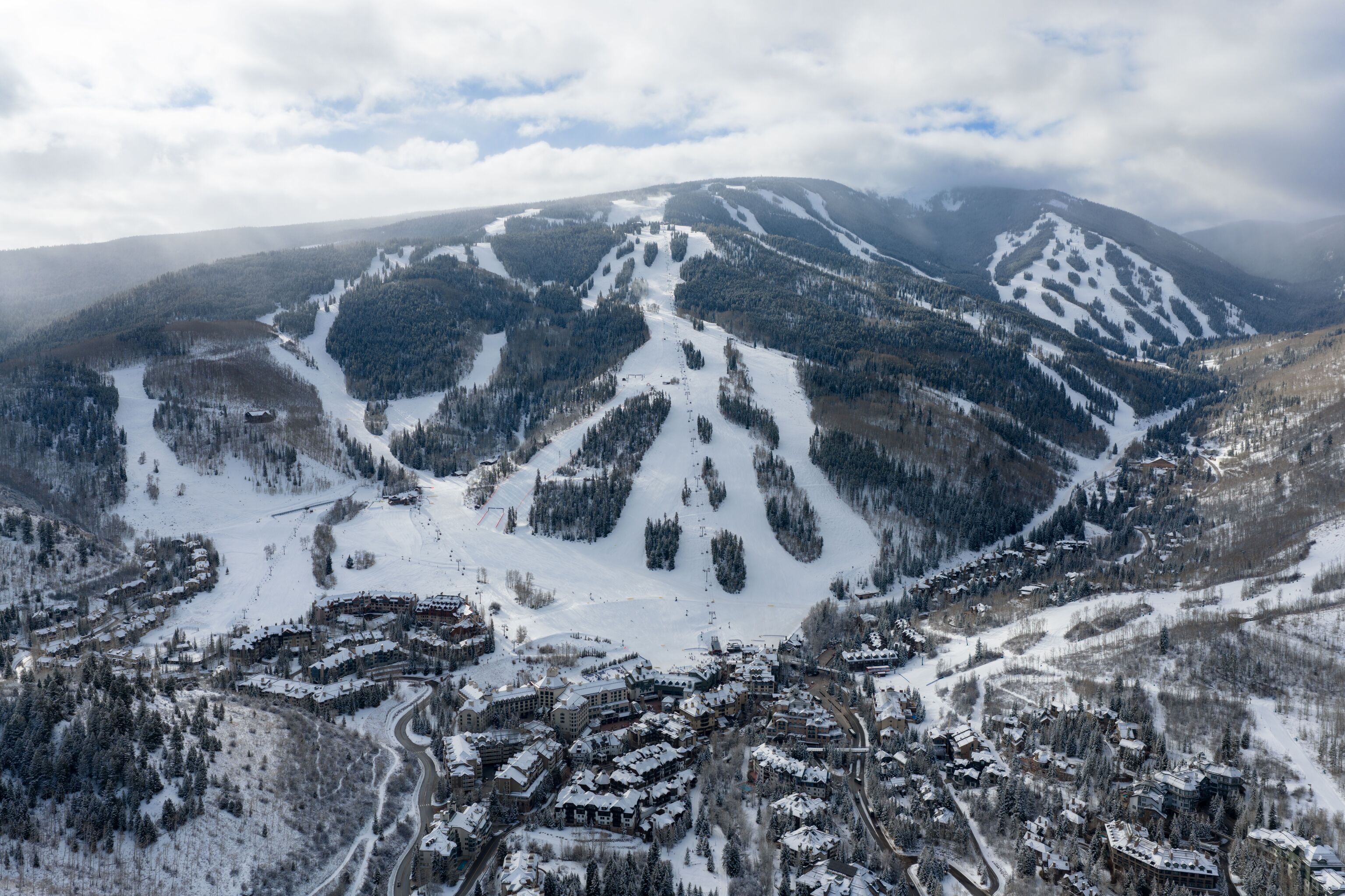 This stunning aerial view showcases a vibrant mountain resort nestled at the base of expansive, snow-covered ski slopes. The village features dense, clustered architecture with warm-toned buildings that contrast beautifully against the winter landscape, offering immediate access to world-class skiing. The majestic, forested peaks in the background provide a dramatic and picturesque backdrop, making this an ideal location for winter sports enthusiasts and mountain vacationers.