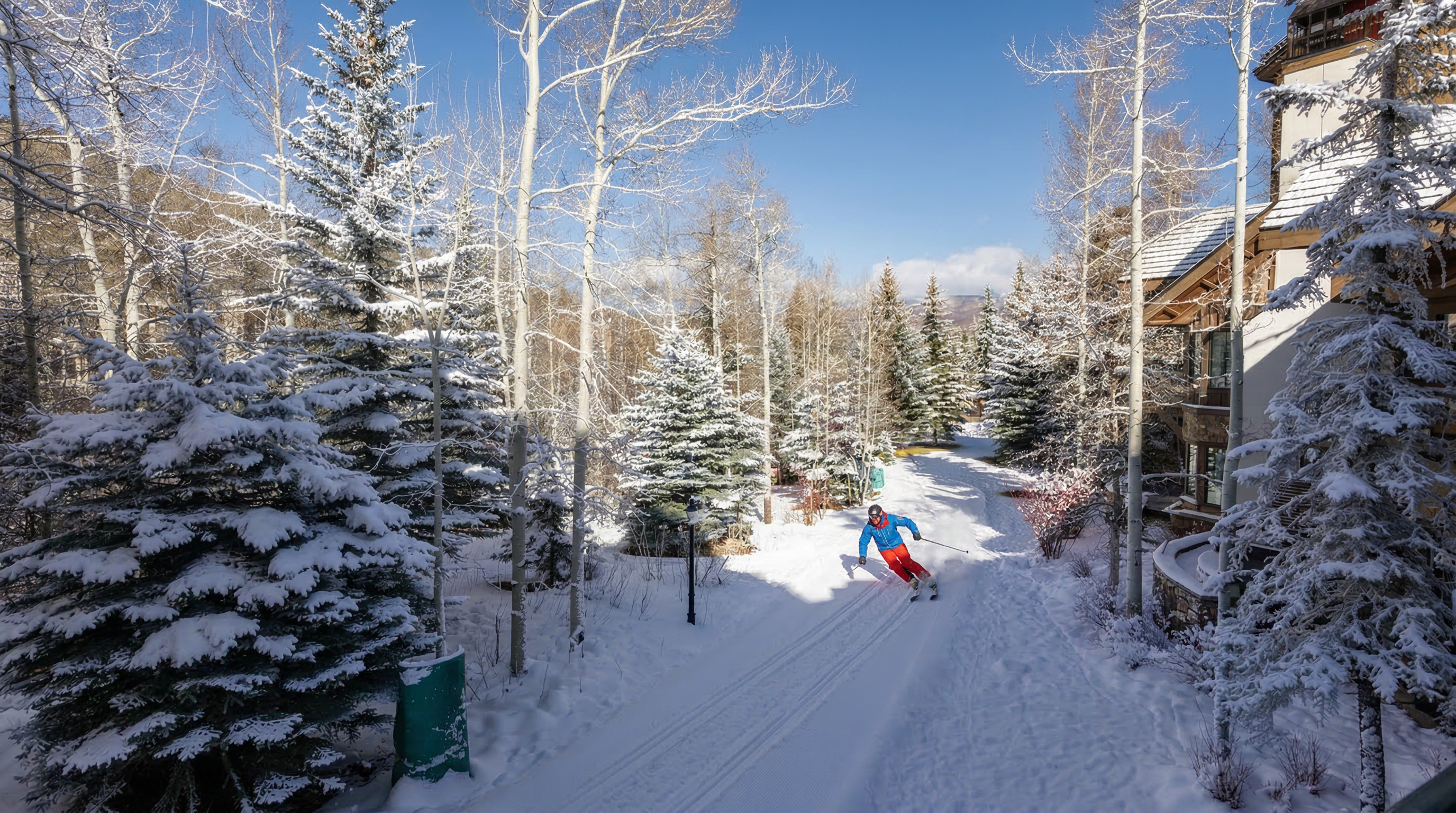 This side view of a mountain property showcases a ski-in/ski-out experience with a groomed trail leading directly past the residence. The building features classic mountain architecture with stone accents and timber details, beautifully nestled amidst a snow-covered landscape of evergreen and aspen trees. The scene captures an inviting alpine atmosphere typical of high-end ski resort real estate.